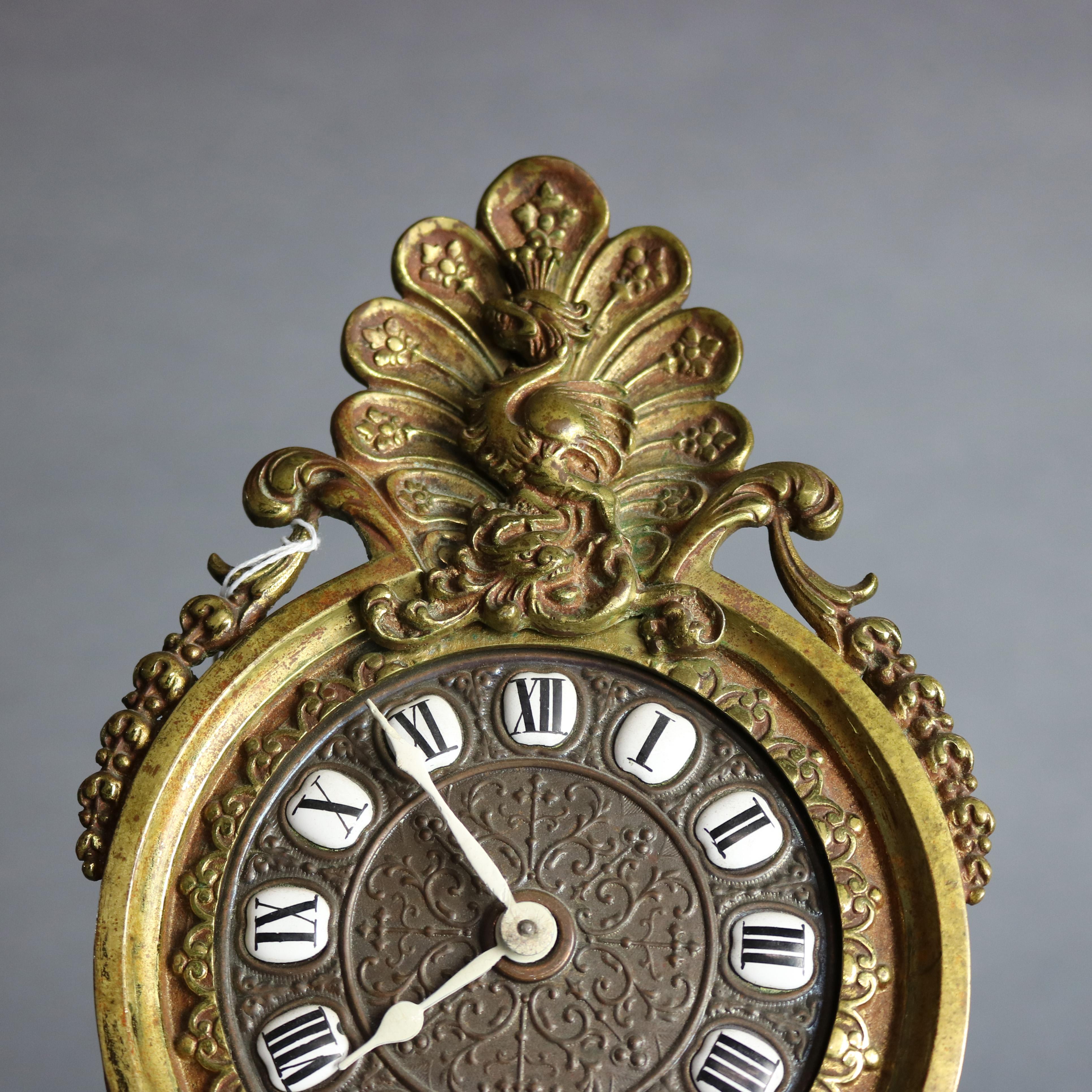 An antique Victorian figural dresser clock offers cast brass construction with peacock finial having flanking cornucopias with inverted bell flowers and surmounting clock face with Roman numerals over central mask with flanking winged dragons and
