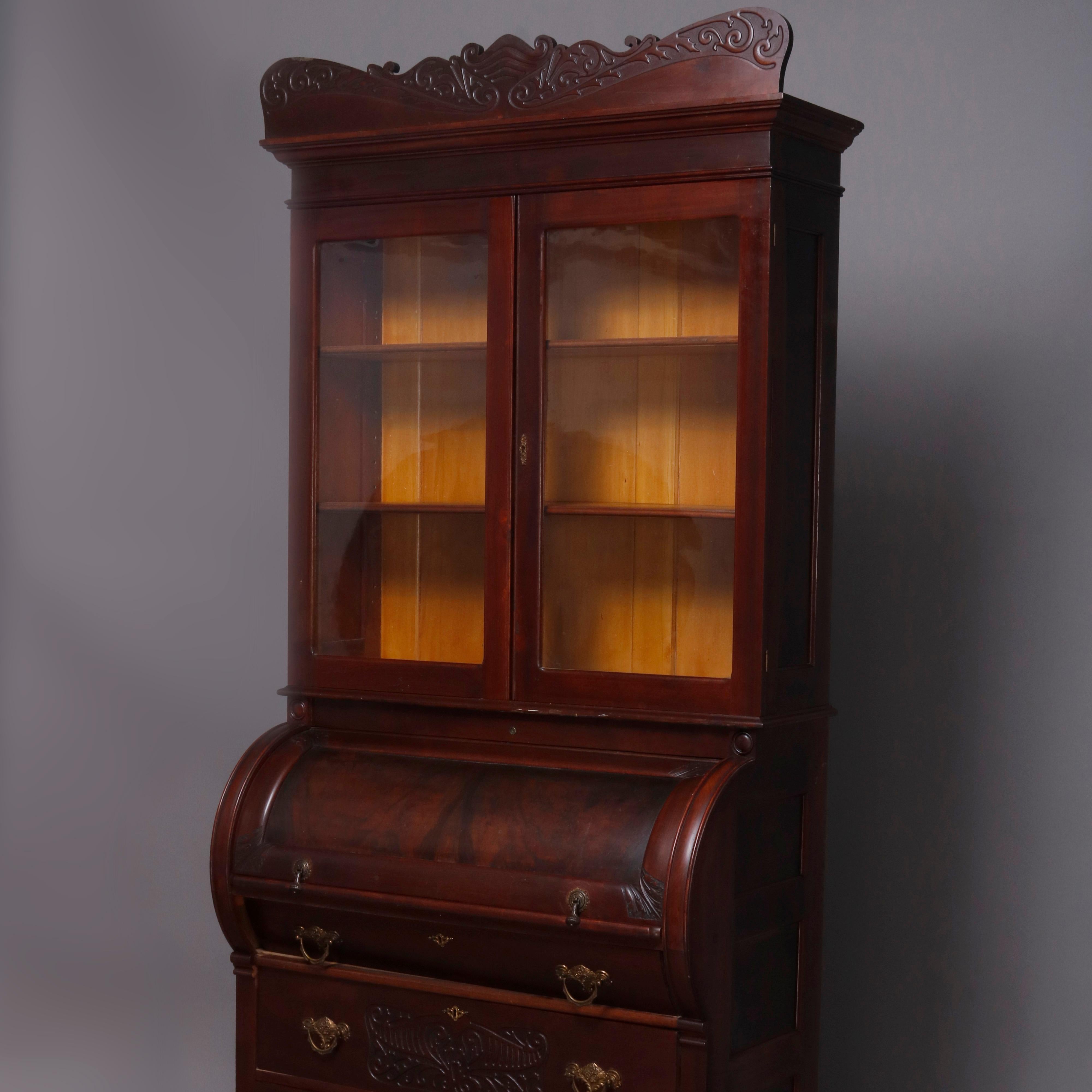 A large antique Victorian cylinder secretary offers carved foliate crown on enclosed bookcase with double glass doors surmounting cylinder desk opening to pull out writing surface with pigeon holes above three long drawers having cast bronze pulls,