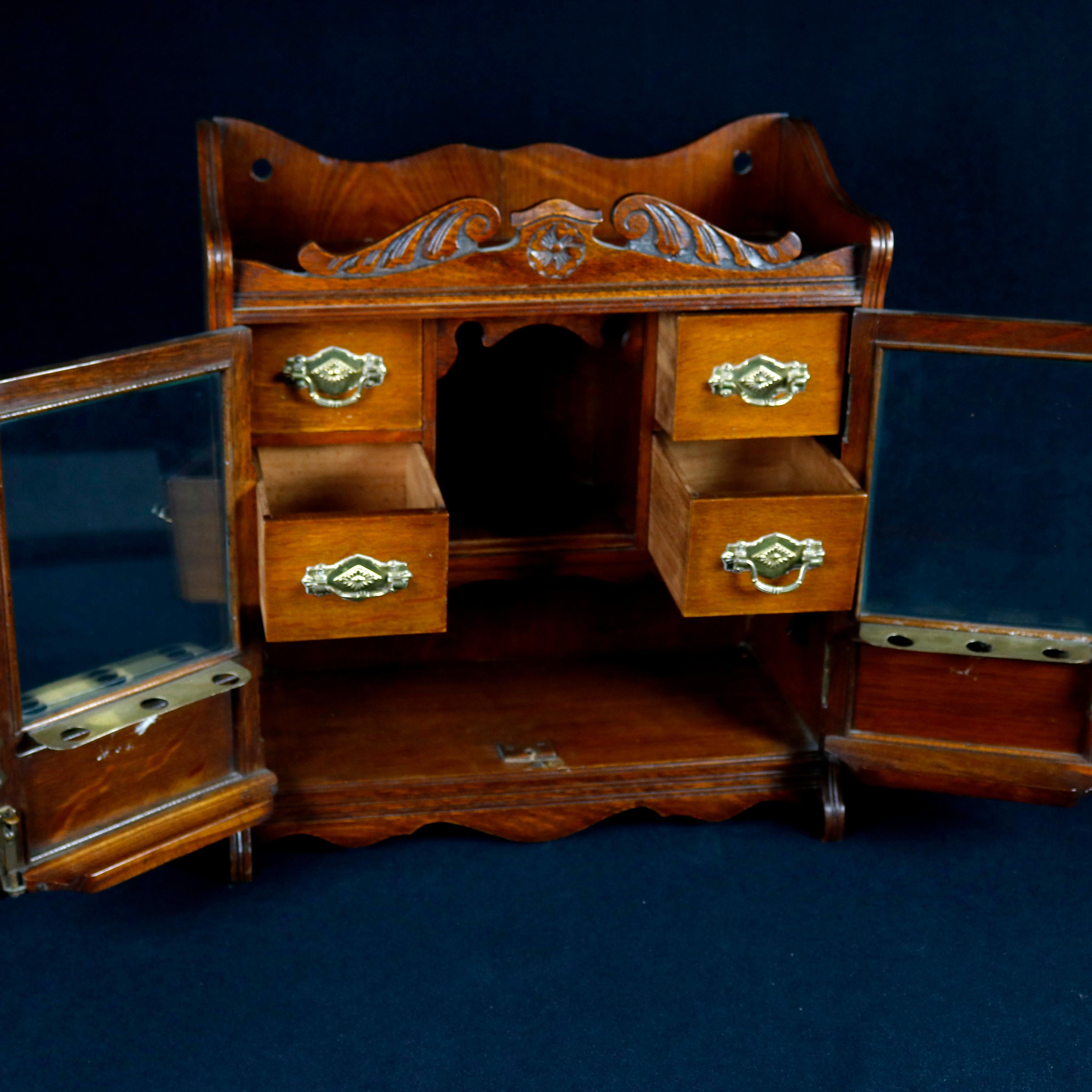 An antique Victorian table top tobacco humidor offers oak construction with foliate carved crest having broken arch pediment surmounting double glass doors opening to reveal interior storage drawers and compartment, cast brass hardware throughout,