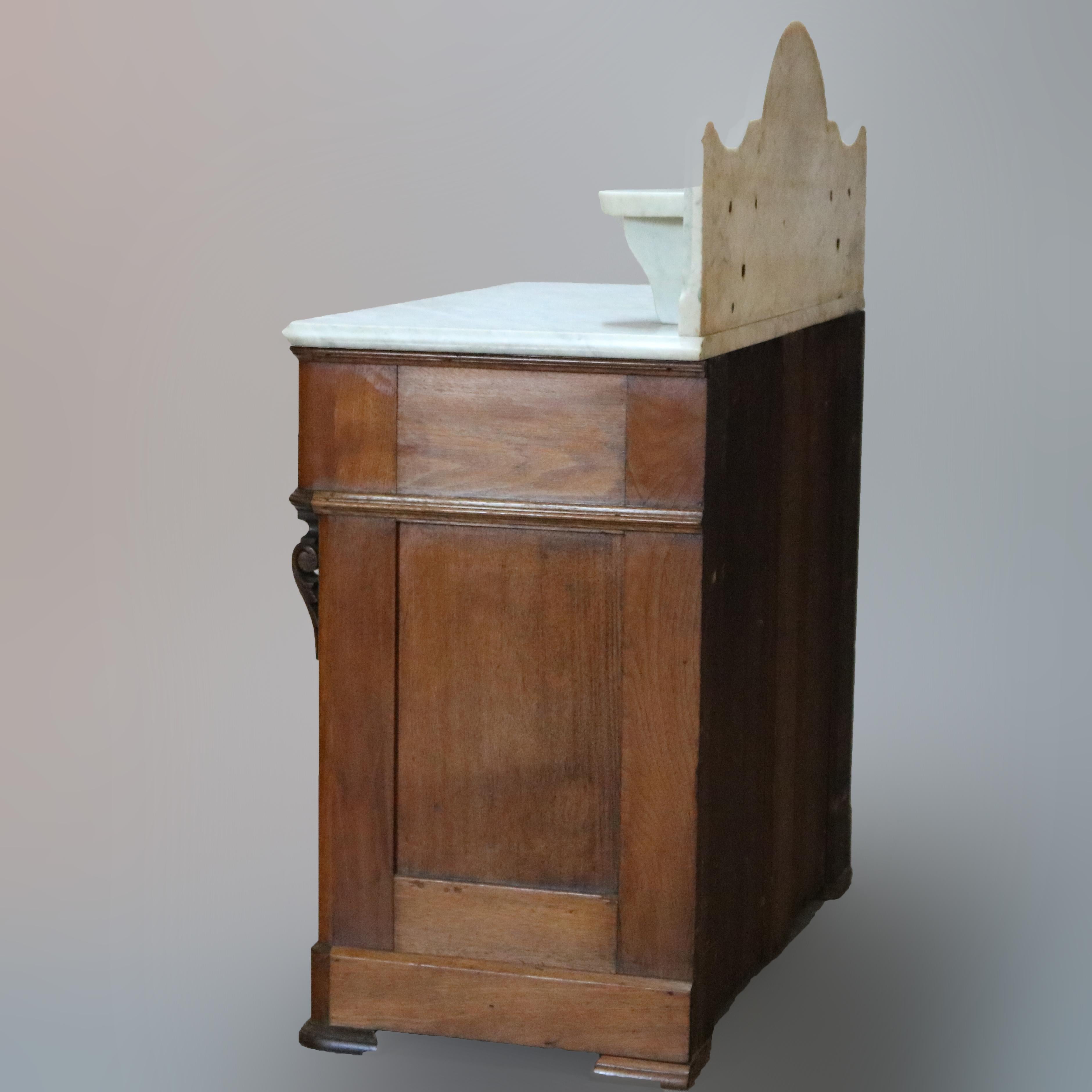 An antique Victorian commode offers shaped marble backsplash with flanking candle stands and surmounting walnut case with marble top and frieze drawer with carved foliate elements and cup handles over double door lower cabinet, circa