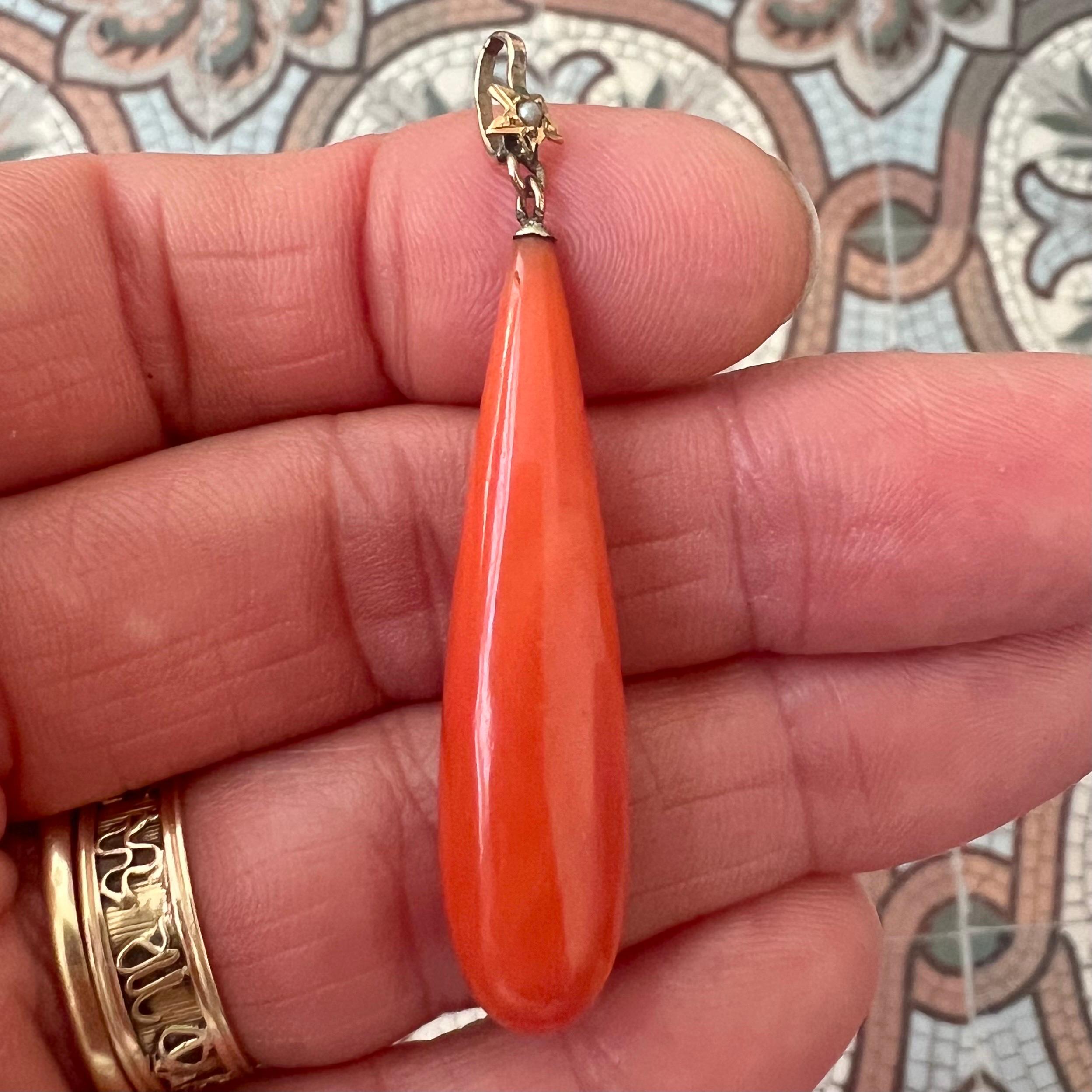 Un bellissimo e grazioso ciondolo antico con una goccia liscia e allungata di corallo naturale del Mediterraneo, sospesa a una delicata cauzione in oro 14 carati ornata da una piccola perla incastonata con un motivo a stella. Il corallo presenta una