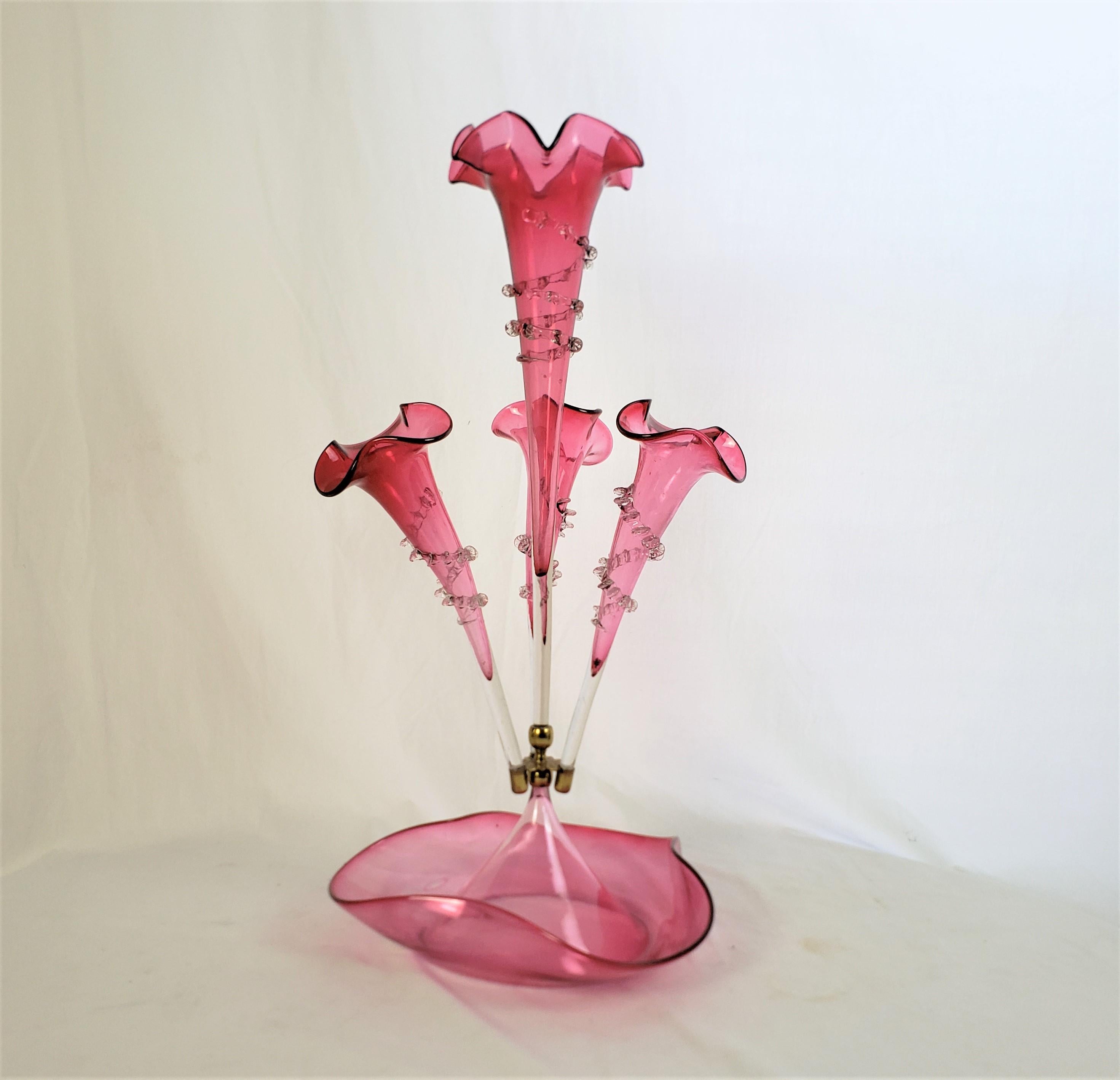 This antique epergne is unsigned, but presumed to have originated from England and date to approximately 1880 and done in the period Victorian style. The epergne is done in a deep cranberry glass with one large central trumpet vase which screws into