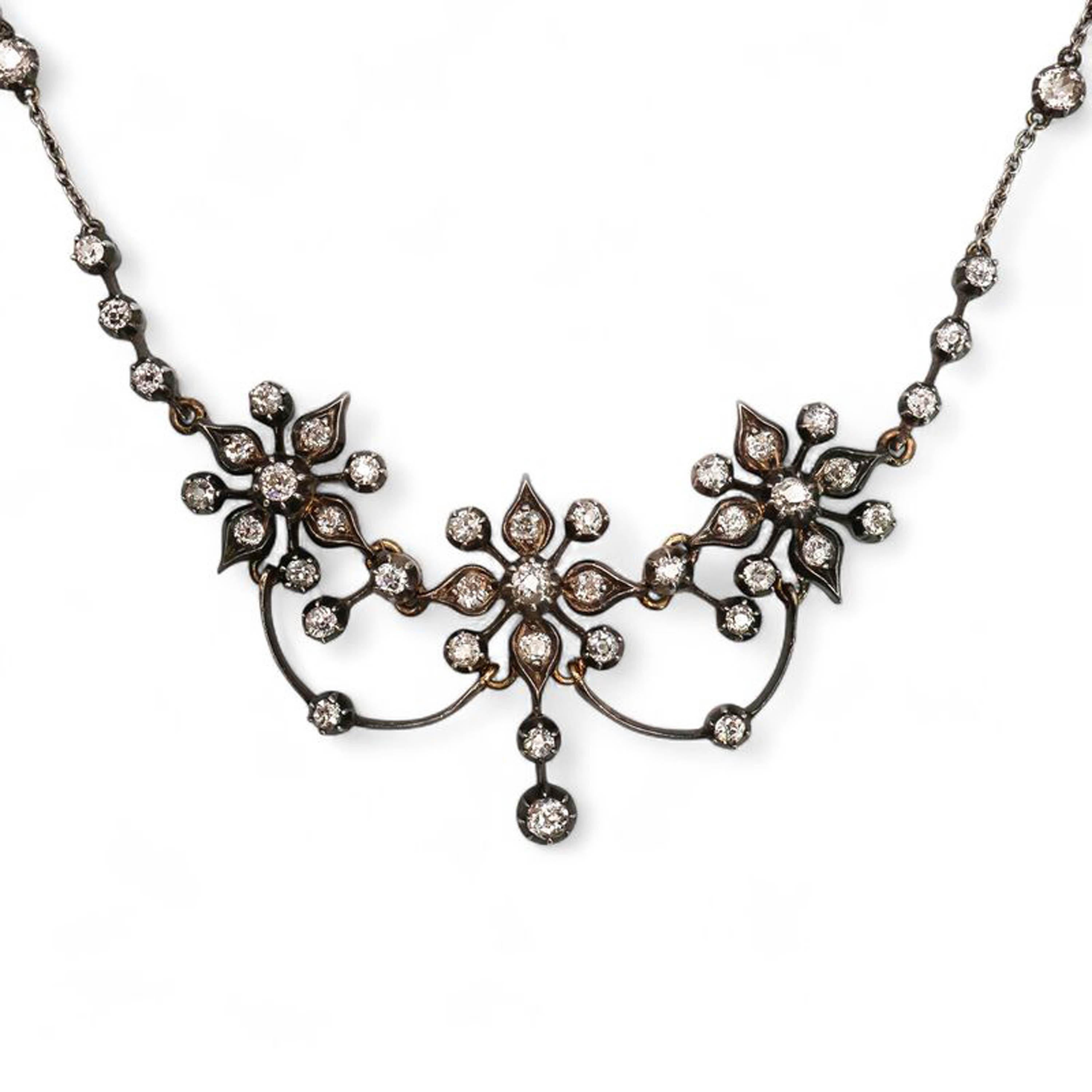 A Victorian diamond, silver and gold flower necklace, featuring an old-cut diamond and cut down set centrepiece of three flowers, at the front, with four, grain set, petals and diamonds on stalks between, the flowers are separated by two knife edge