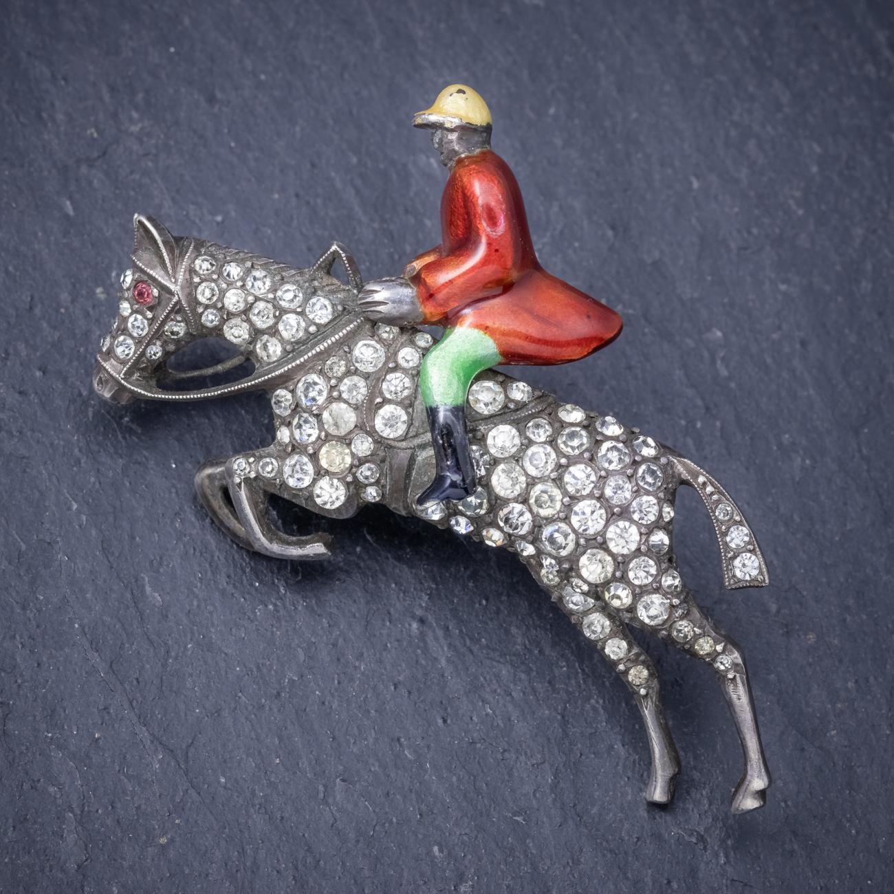 A wonderful antique Victorian riding brooch depicting a horse and rider leaping over an imagined fence. The rider is detailed with a red Enamel jacket, yellow helmet and green jodhpurs. The horse is decorated with white Paste stones which sparkle
