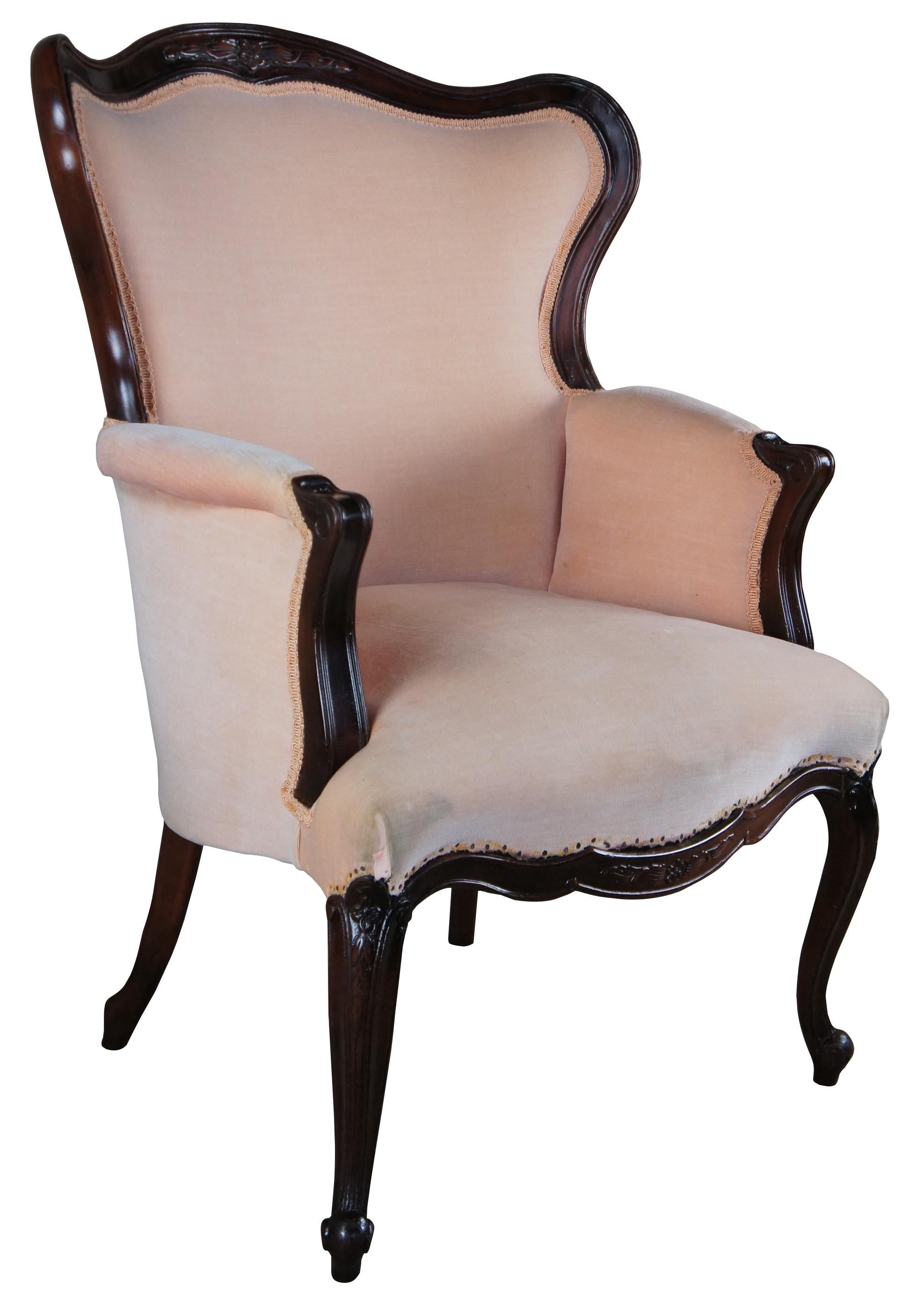 Early 20th century Late Victorian French Provincial inspired wingback arm chair. Made from mahogany with a serpentine carved crest and lower apron, pink fabric and cabriole legs with knee carved foliate leading to scrolled feet. 
 