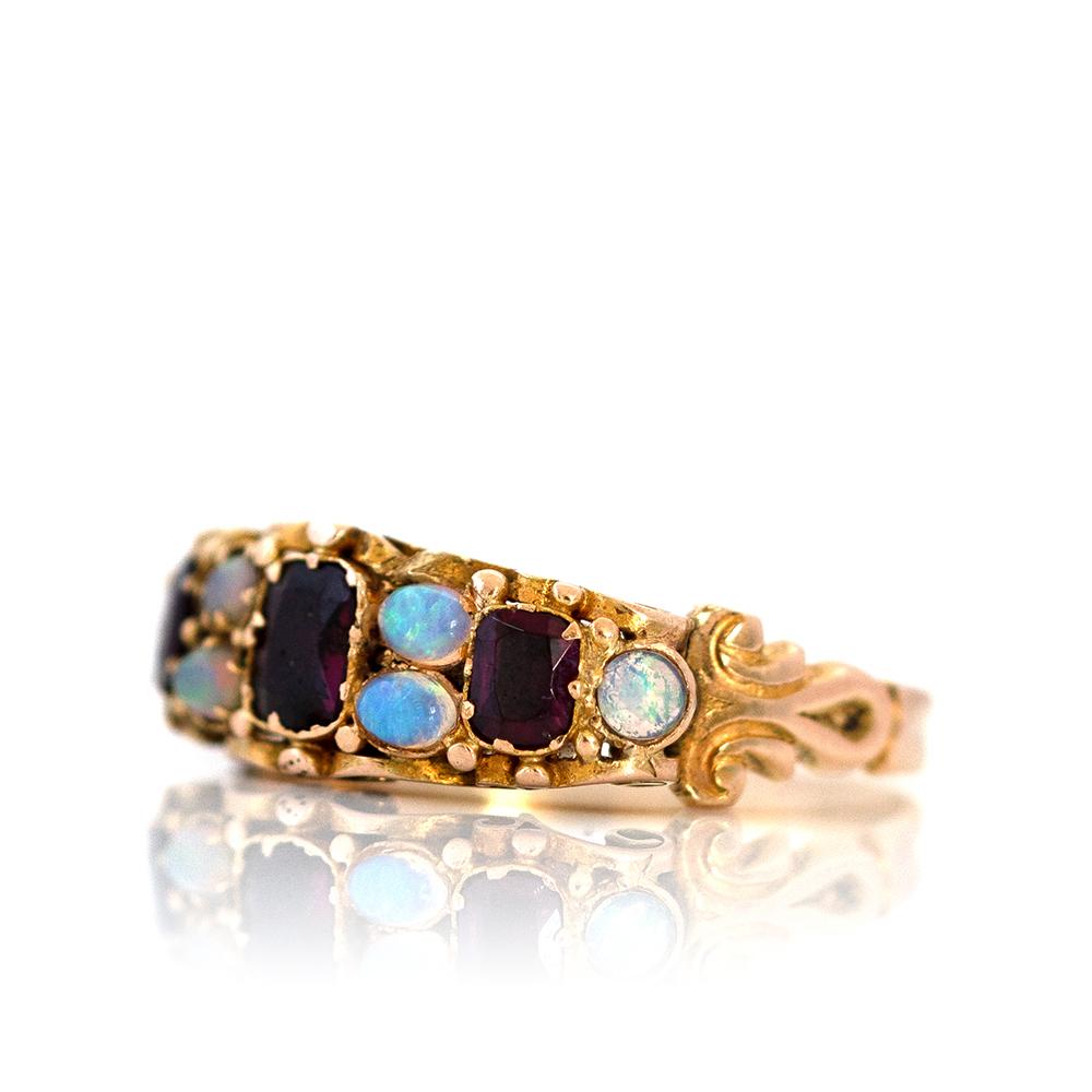 This richly detailed Victorian garnet and opal ring is a captivating example of 19th-century  (c. 1860s–1880s) sentiment and craftsmanship. Set in warm 15ct yellow gold (unmarked but tested), the ring features a striking trio of deep red garnets,