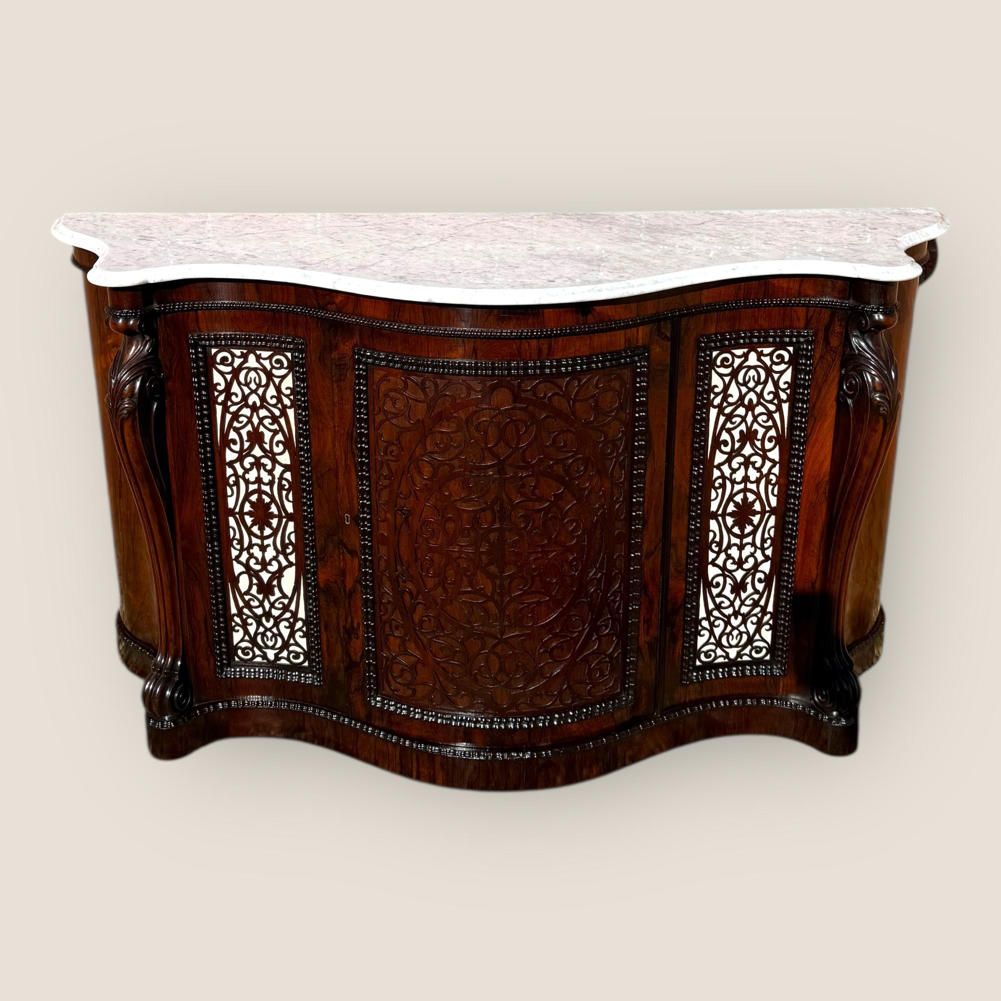 A Victorian rosewood credenza of serpentine form, circa 1874, attributed to Gillows of Lancaster. The top is inset with a white Italian Carrara marble slab, veined in grey. The central cupboard door is overlaid with intricate fretwork framed by