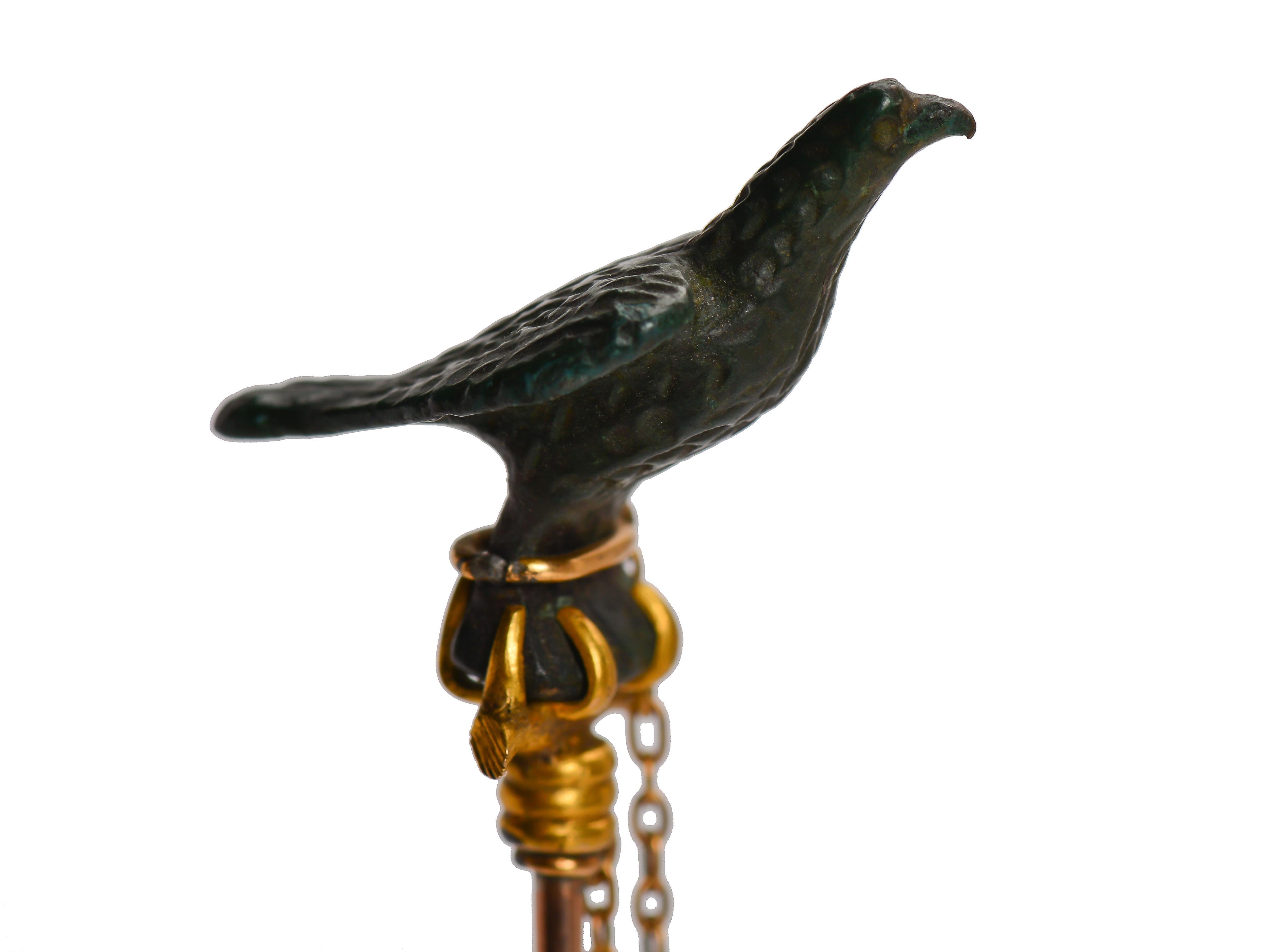 This one of a kind, Victorian stickpin shows a falcon immortalized in bronze and gold. The bird of prey perches, chained on his bar in an alert, mighty pose. The bronze has a slight green patina. The top perch and chain are 20Kt gold. The pin is 9Kt
