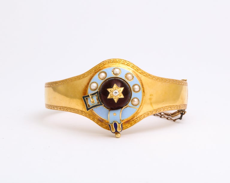 A Victorian bracelet in 15 Kt gold has a large dome cabochon garnet in the center that is set with a gold star and small diamond. A border of light blue enamel surrounds the garnet and that is set with natural pearls. A deep blue buckle and strap