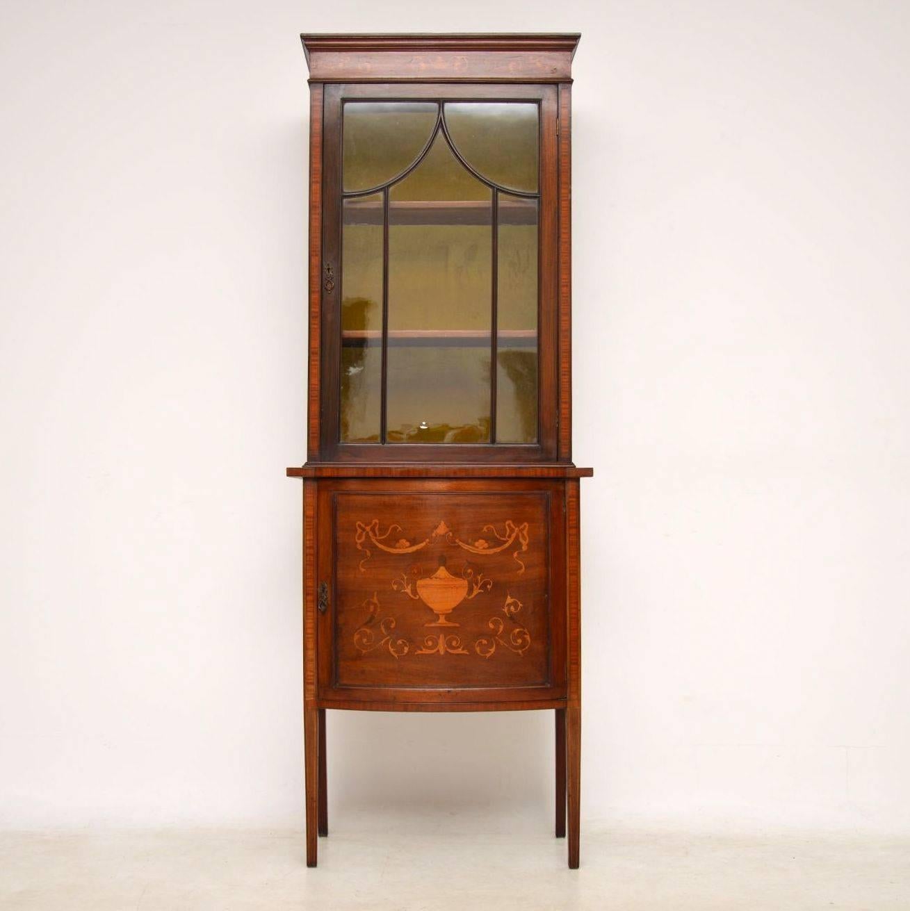 This slim antique mahogany and satinwood display cabinet dates circa 1890s period and is in good condition. The top half is a glazed cabinet lined inside. It sits on a bow fronted cupboard with beautiful inlays and stands on tall legs. Please