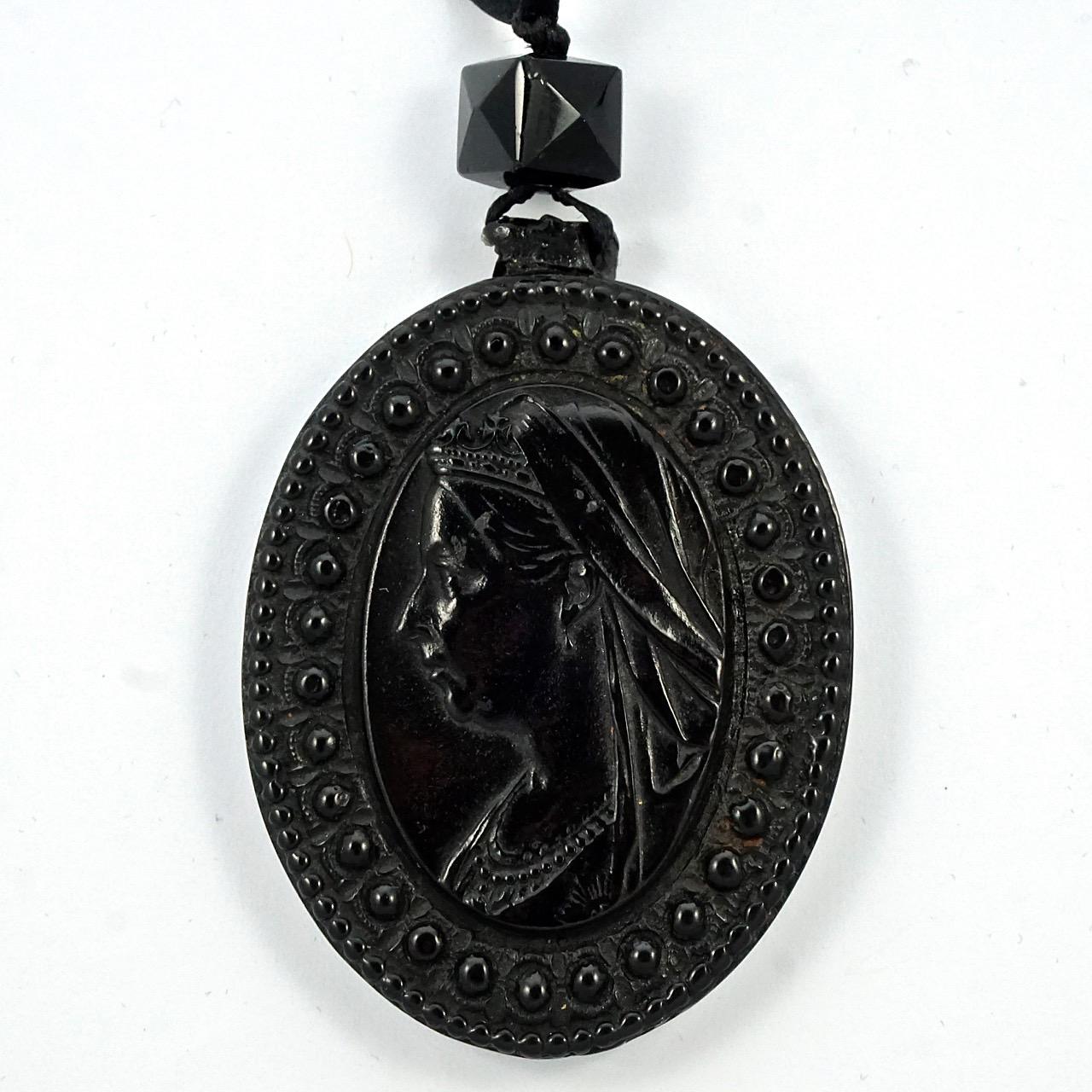 Antique Victorian large faux jet moulded pendant, hung on black ribbon with a pinchbeck dog clip. On the front is a profile of Queen Victoria in relief with a decorative edging, and the back is inscribed 'May Saints Embrace Thee with a Love like
