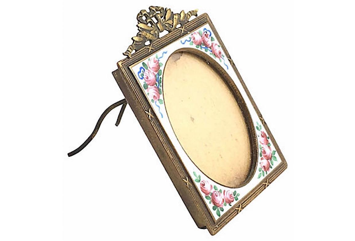 Miniature Victorian bronze picture frame featuring a bow on top and white enamel decorated with pink flowers on each corner. Oval picture opening measures approximately 1
