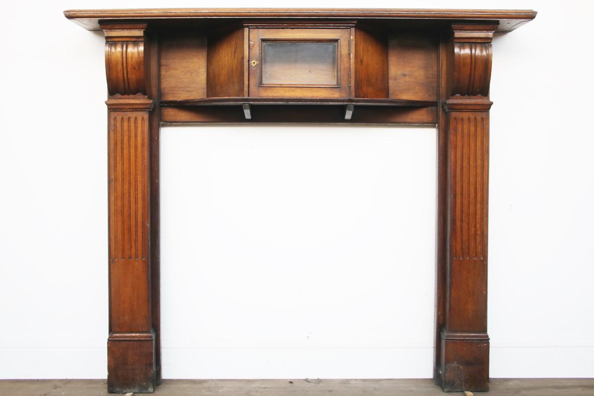 Antique Victorian oak fireplace surround. Carved end brackets support the generous mantle shelf and sit above fluted jambs. The frieze is centred with a central compartment complete with original glass.

In as found condition with expected signs