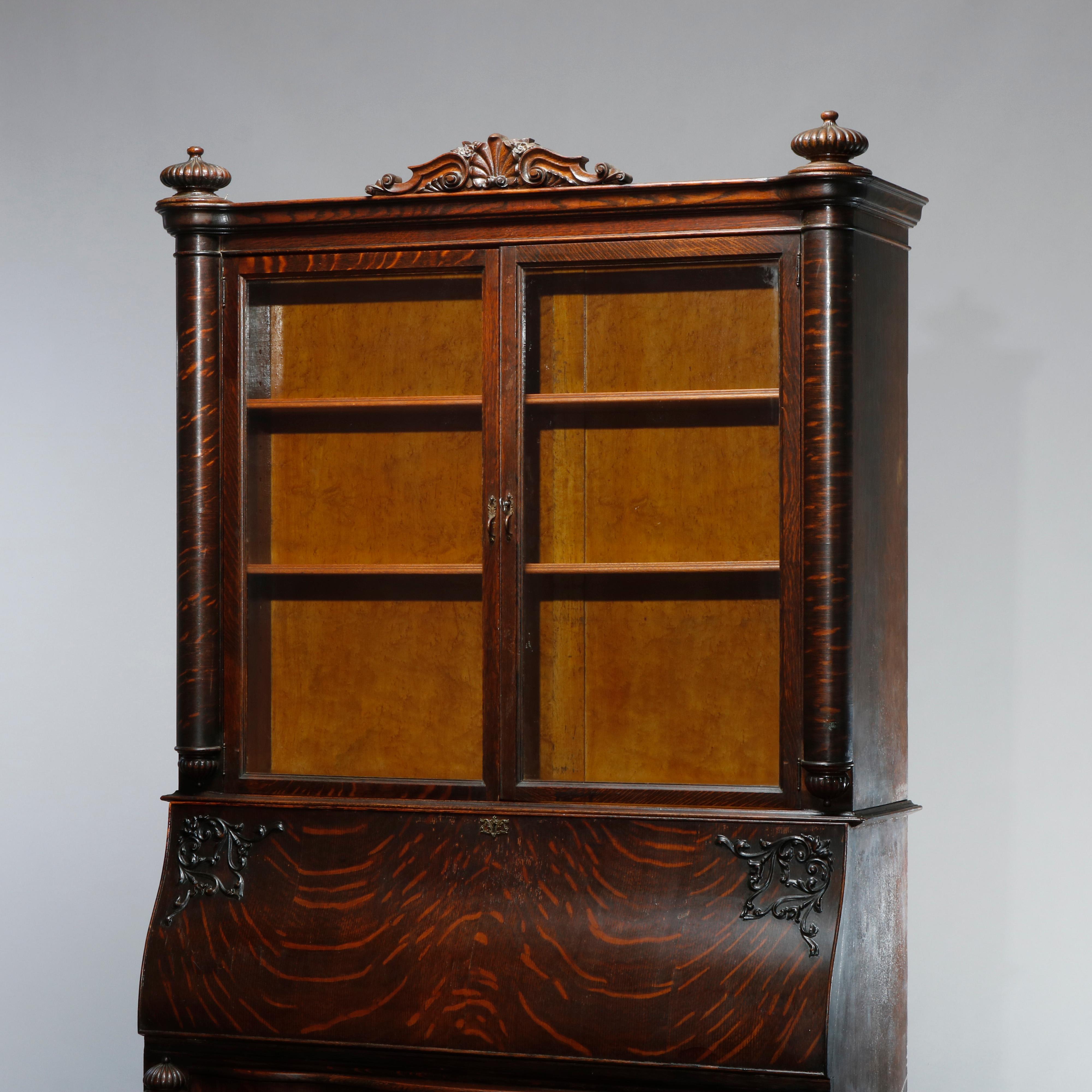 An antique secretary in the manner of RJ Horner offers quarter sawn oak construction with upper case having carved and pierced foliate crest flanked by melon bun finials over double glass doors opening to shelved interior; lower with drop front desk