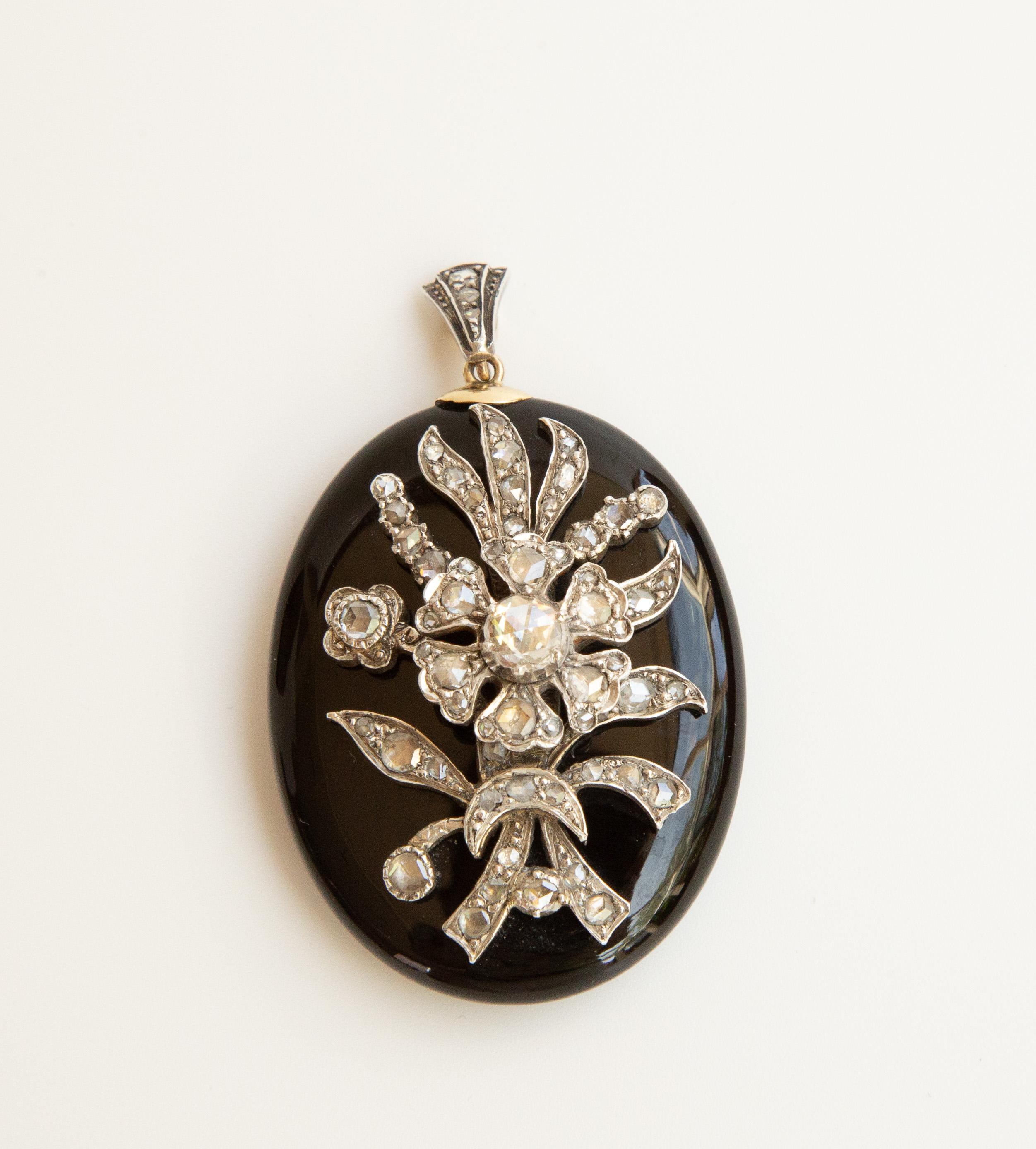 An antique Victorian oval onyx locket pendant with a silver flower applique set with rose diamonds on  foil. The pendant features on the back side a locket to store a mini photograph or a tiny item. The locket closes with a plastic cover (likely