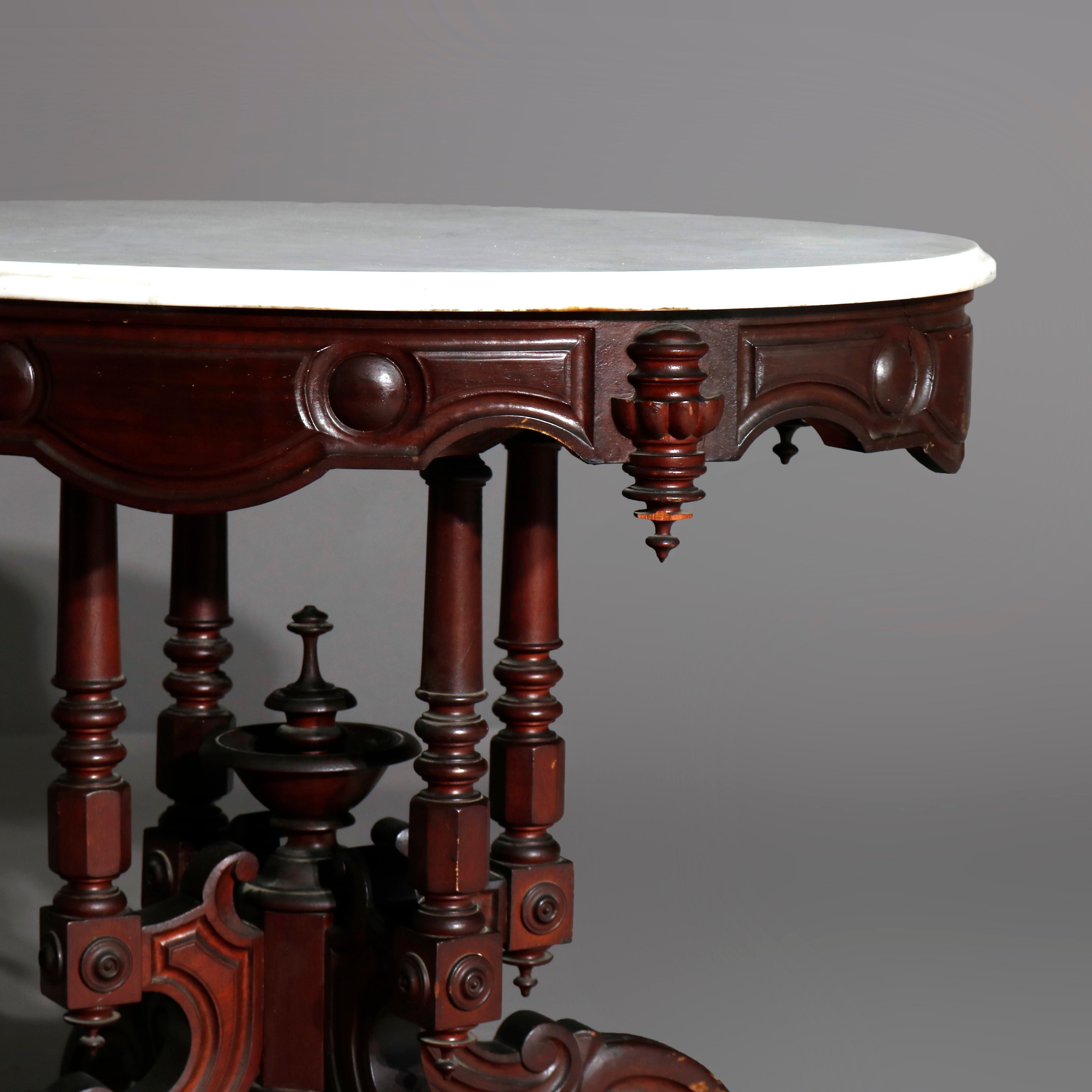 An antique Victorian parlor table offers an oval and beveled marble top surmounting a walnut base having shaped skirt and drop finials over four turned columns and carved urn form center finial, raised on carved scroll form legs with casters, circa