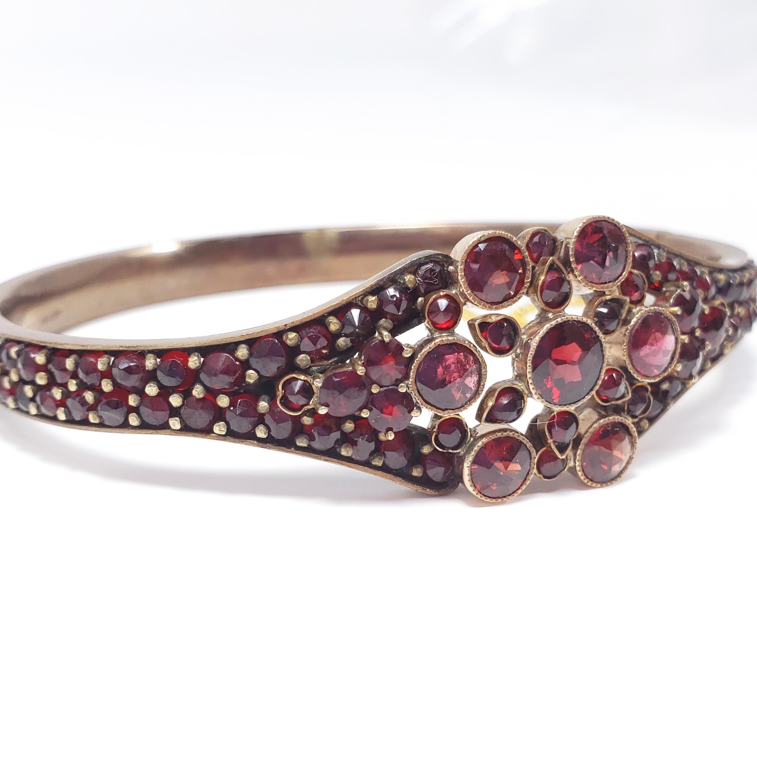 An antique bracelet decorated with pave garnets. The garnets on the face are open back and set in sterling silver, aligned in a decorative pattern, while the gemstones around the band are prong set. Fastened with a box-style clasp and safety