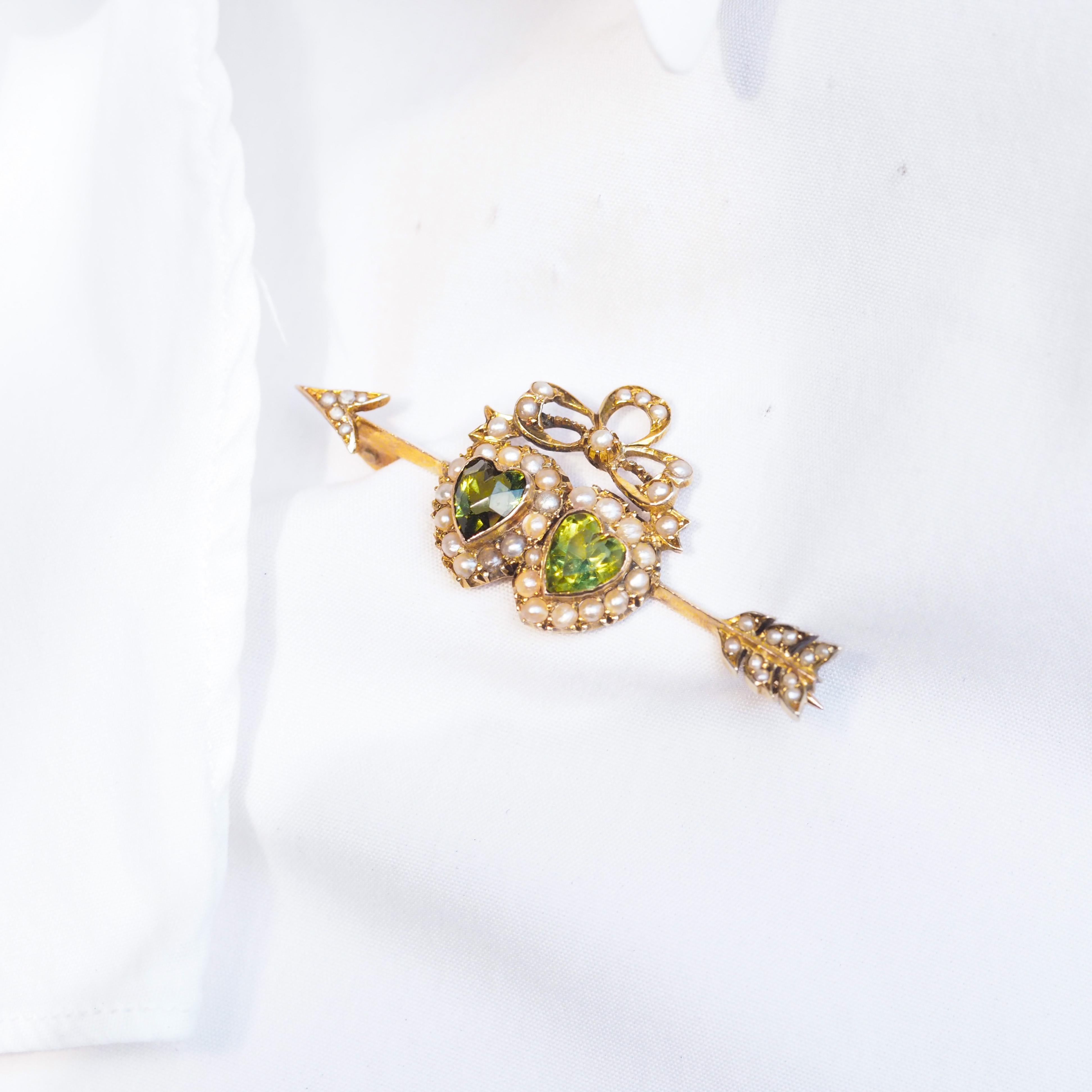 Estamos encantados de ofrecer este encantador broche victoriano antiguo de doble corazón de peridoto fabricado hacia 1900.

El broche está maravillosamente realizado en oro de 9 ct en forma de flecha que atraviesa dos corazones. La sección central
