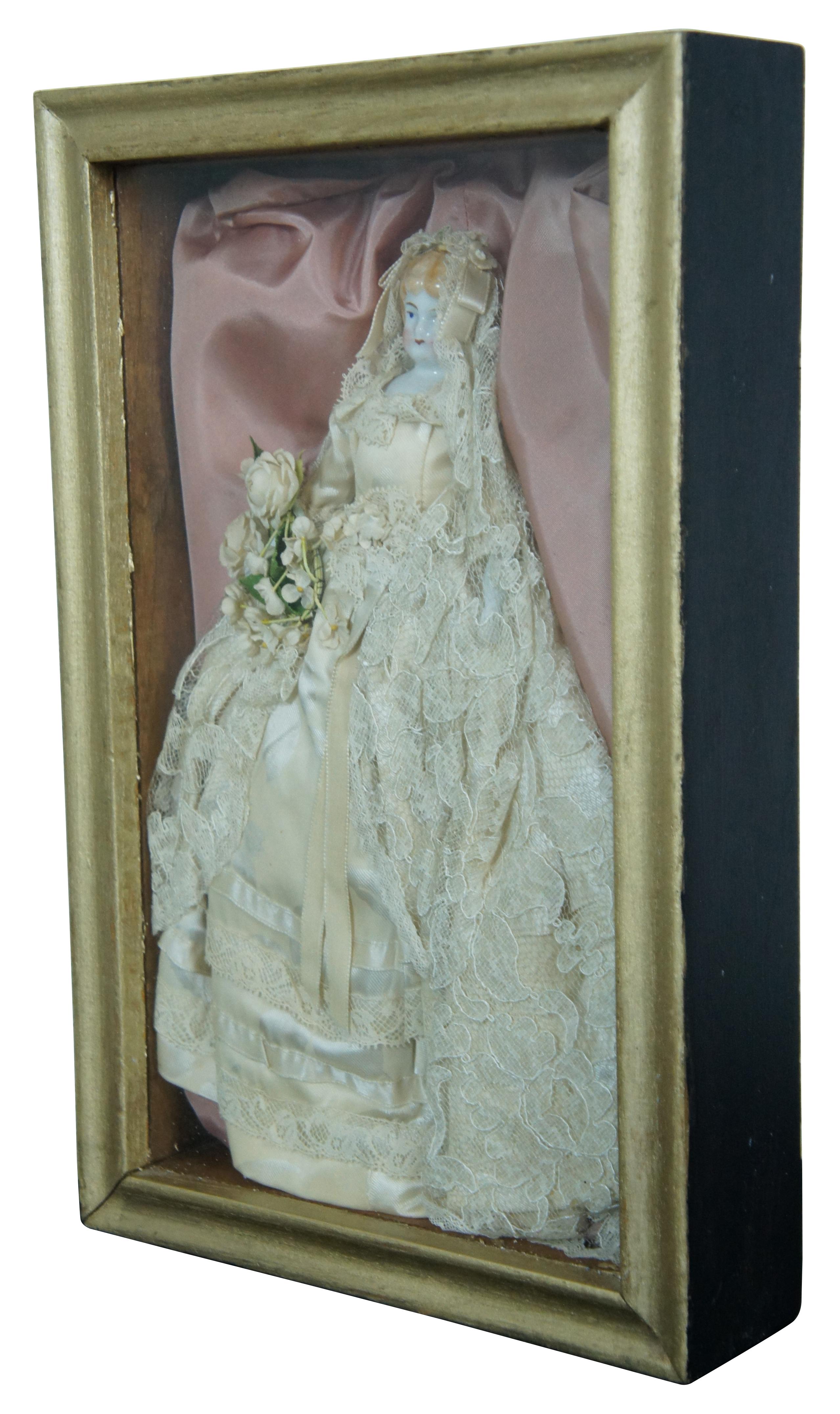 Antique mid 19th century molded head porcelain doll with blonde hair and blue eyes, dressed in a lovely floral wedding gown with lace over-skirt and bouquet of flowers, displayed in a pink padded shadowbox with gilded frame.
 