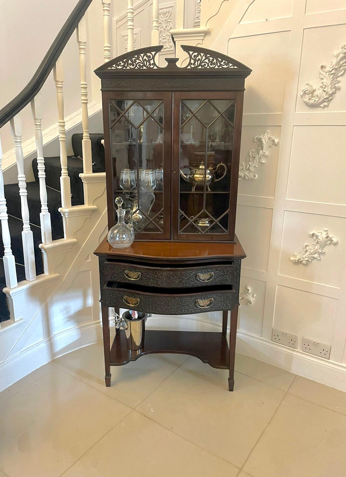 Antique Victorian quality carved mahogany display cabinet having a quality carved pediment above a pair of astral glazed doors opening to reveal two adjustable display shelves supported by a serpentine shaped two drawer carved mahogany table with
