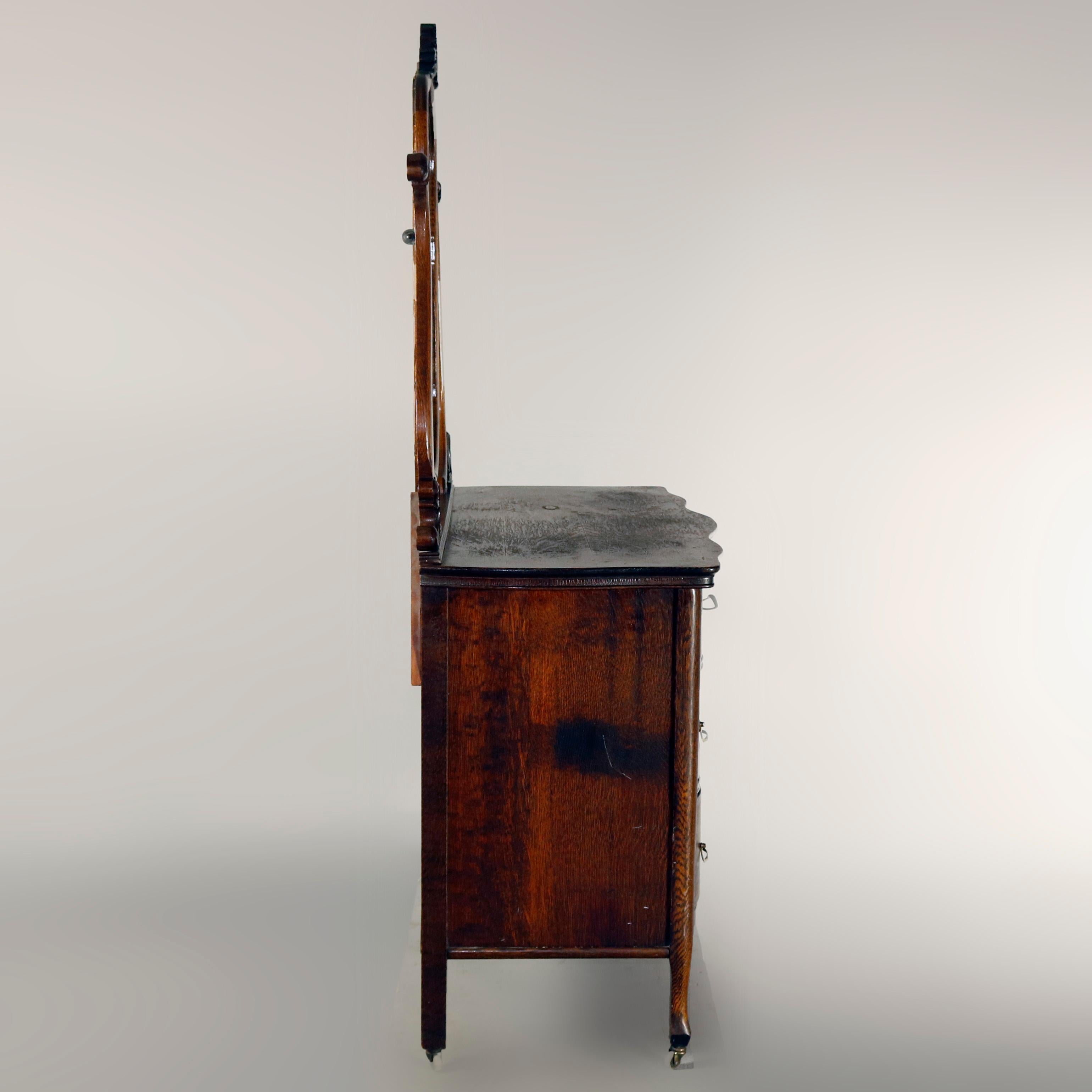 An antique Victorian chest of drawers features quarter sawn oak construction in serpentine form with two smaller drawers over two long drawers surmounted by shield shape mirror with carved foliate crest and raised on s-scroll arms with carved