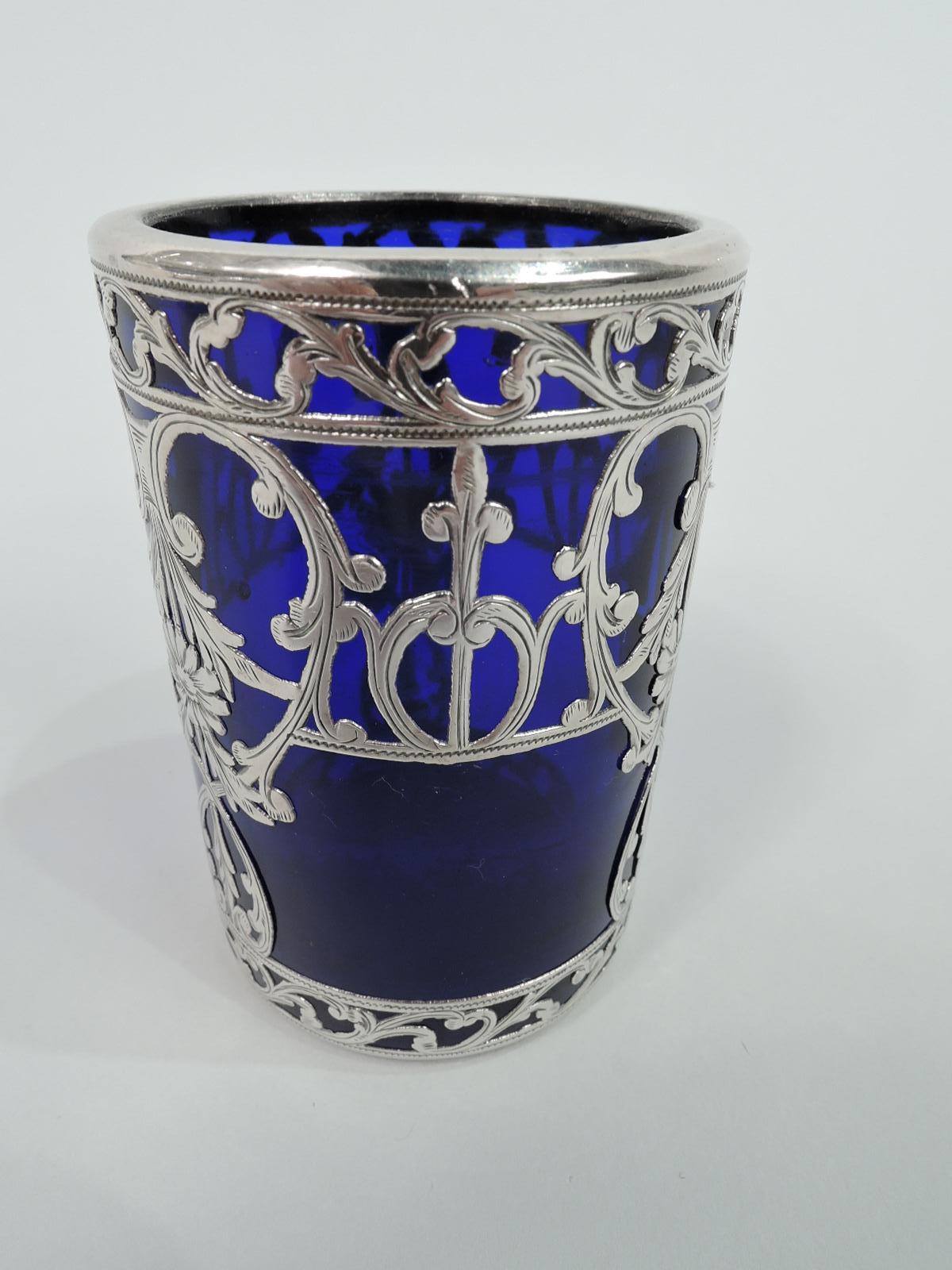 Turn-of-the-century glass bud vase with engraved silver overlay. Cylindrical beaker with flat rim in silver collar. Overlay patter comprising leafing scrolls and flowers between leafing-scroll borders. Glass is cobalt blue.