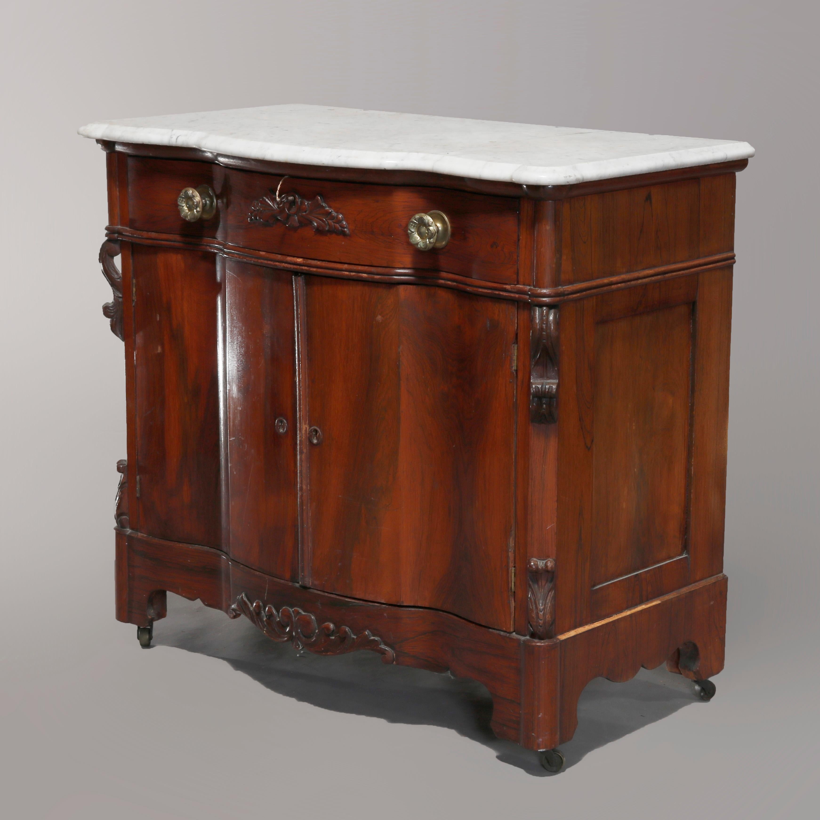 An antique Victorian Rococo commode offers rosewood construction in serpentine form with shaped marble top surmounting base with frieze drawer over double door lower cabinet, carved acanthus and foliate elements throughout, circa 1860.

Measures: