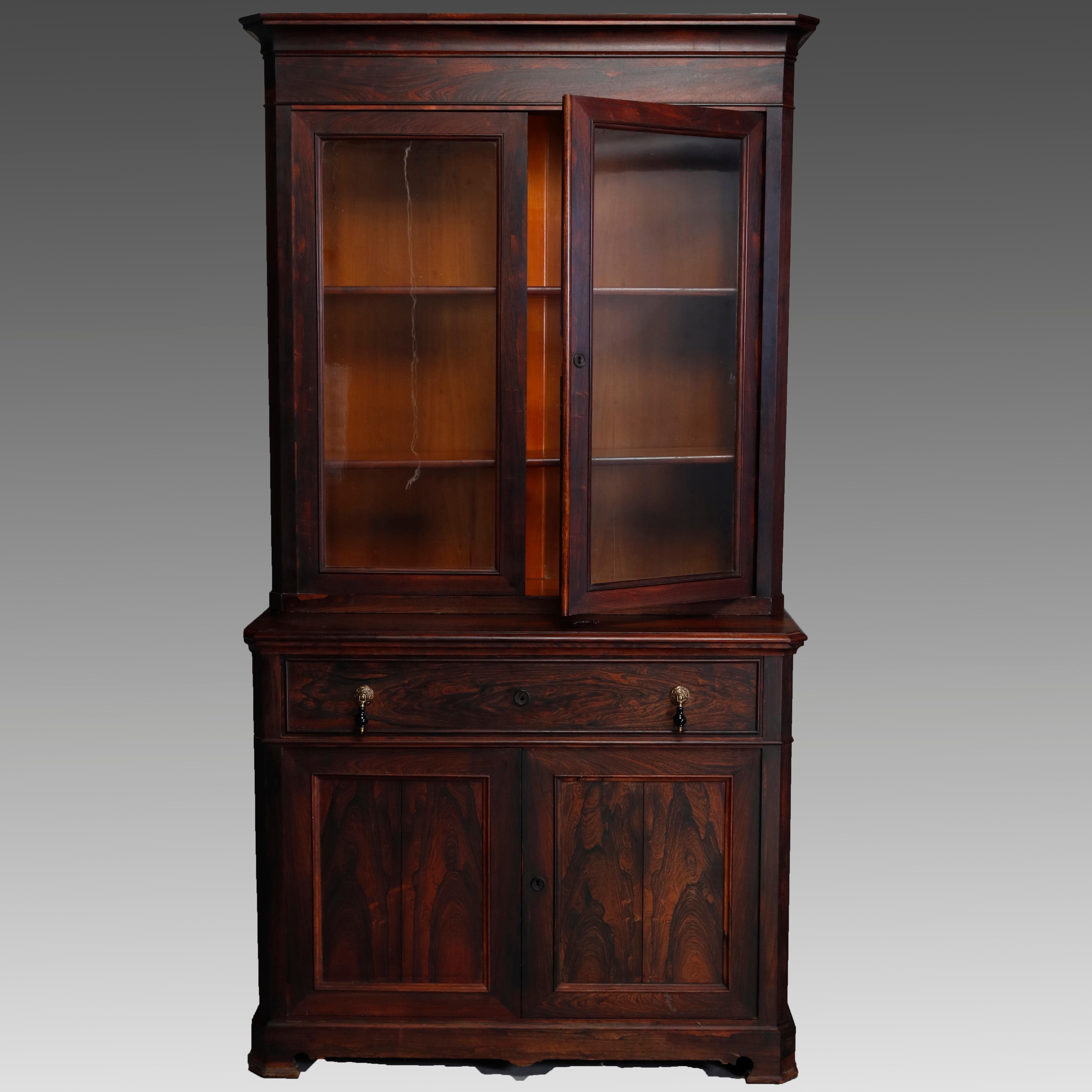 An antique Victorian secretary offers deeply striated rosewood construction with upper bookcase having double glass doors and surmounting lower case with drop front desk with teardrop pulls opening to reveal writing surface with storage compartment