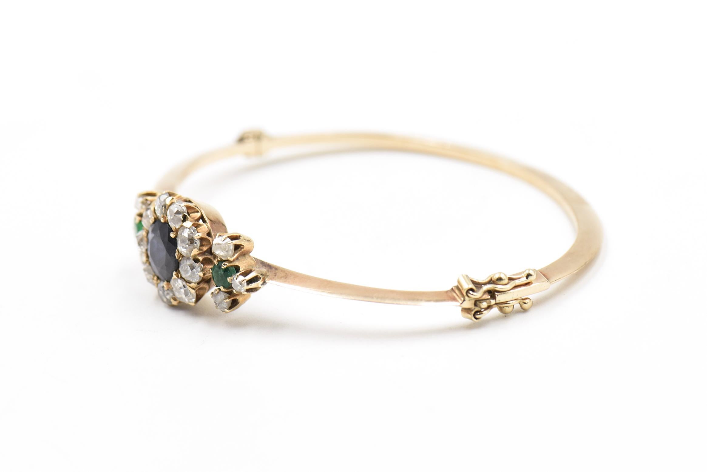 Victorian flower bracelet featuring a faceted sapphire summoned by old mine cut diamonds with emerald and diamond leaf accents on the sides.  The design sits on a knife edge 14k yellow gold bangle bracelet.  The bracelet hinges open and has a figure