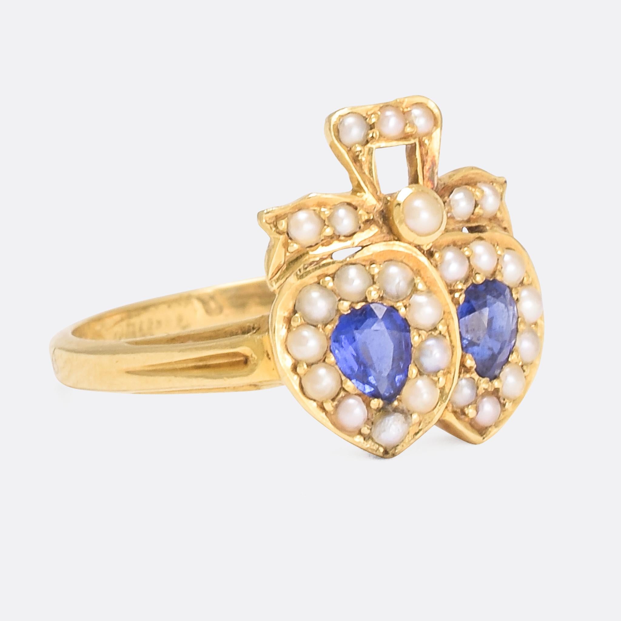 A beautiful antique double heart ring set with vibrant blue sapphires and split pearls. It's a lovely design, the two hearts overlap where they meet, and sit beneath a pearl-set bow that's reminiscent of two swans. Modelled in 18k yellow gold, it