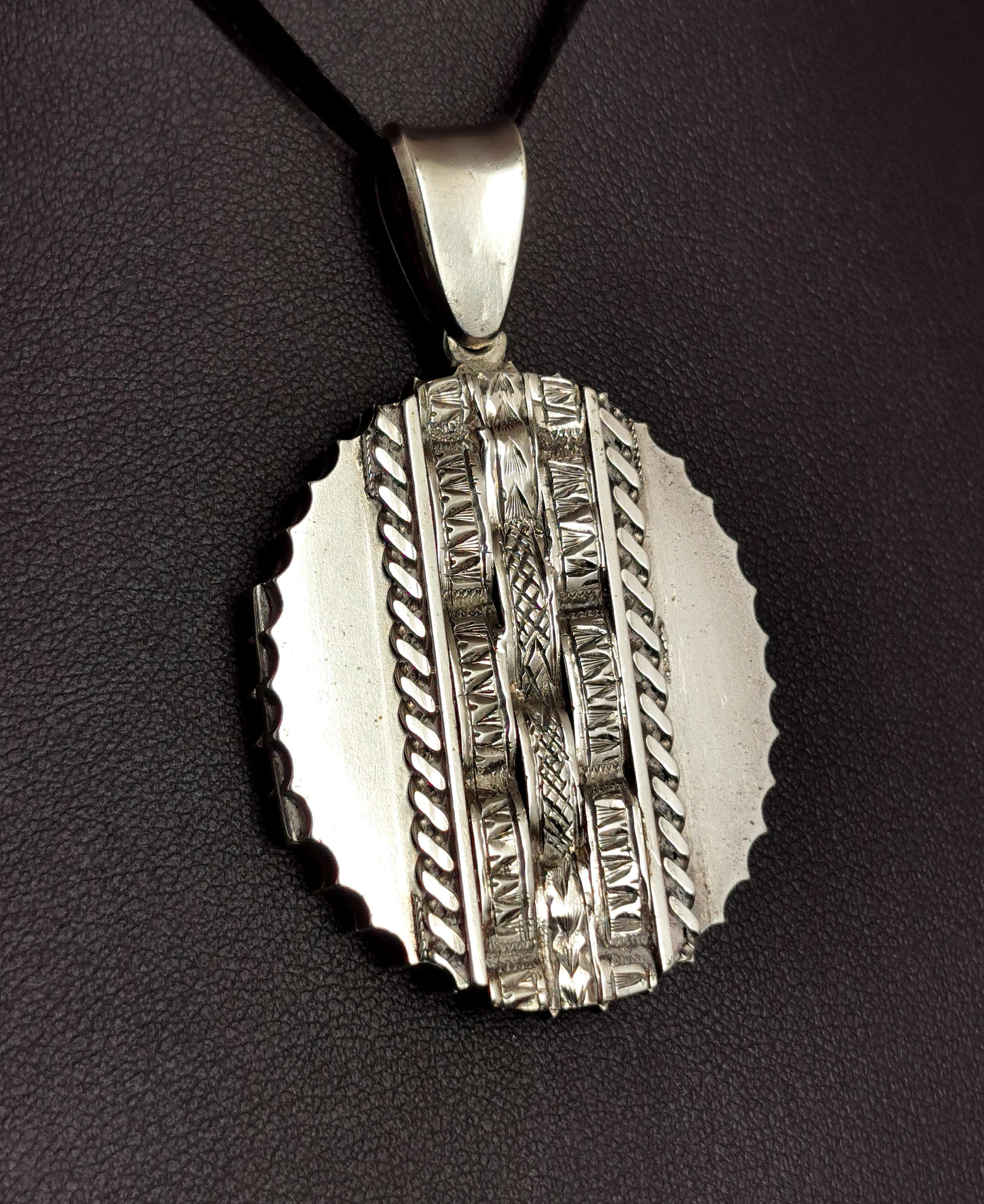A beautiful antique Victorian sterling silver locket pendant.

It is engraved to the front with an unusual applied curved weave design down the front.

It has a large, integral bale and is an oval shape with a wavy zig zag edge.

The reverse of the