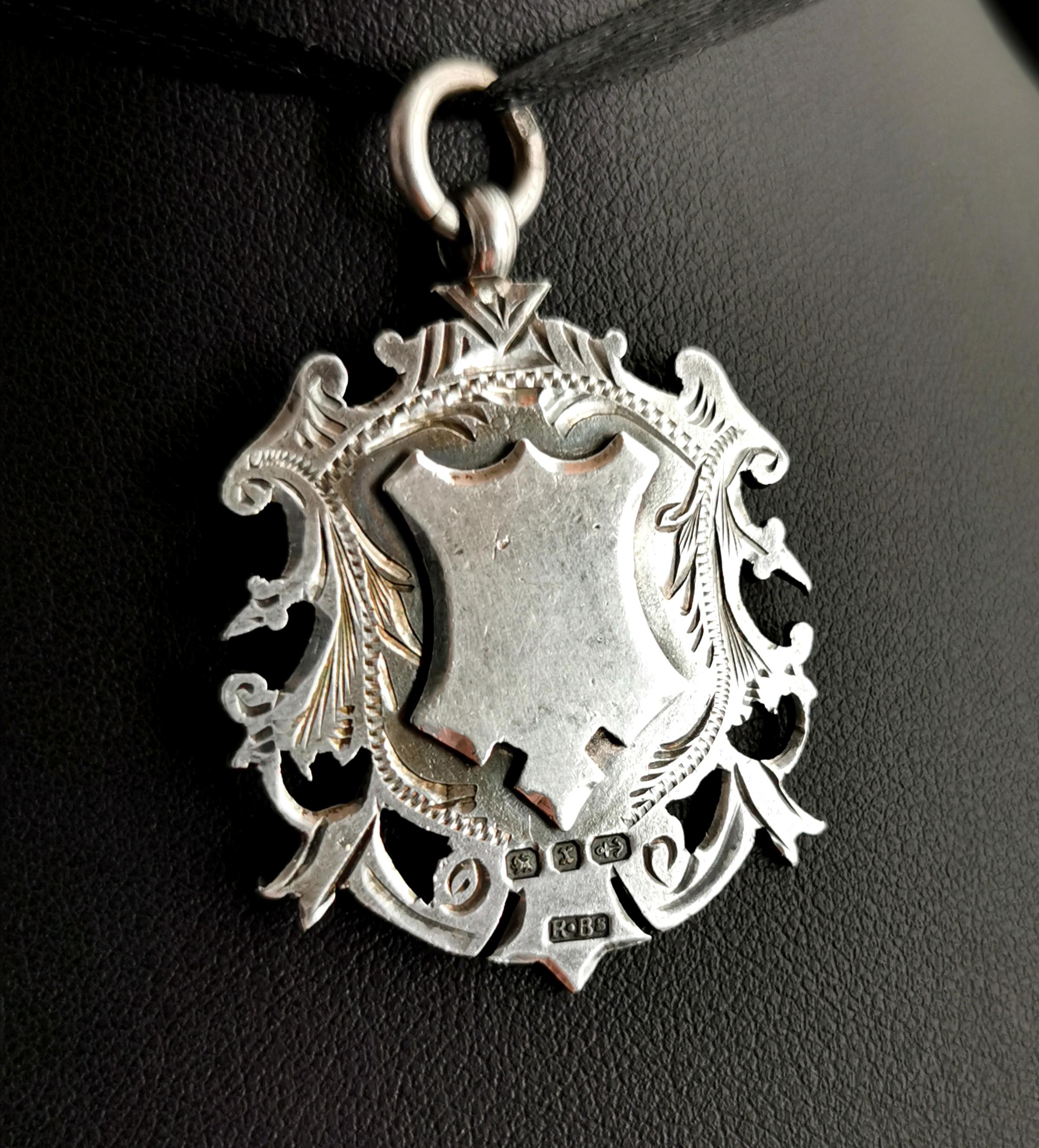 An attractive and elaborate antique sterling silver fob pendant.

A larger fob with a good weight to it, it is a shield shaped piece with a chased, engraved cut out border.

The fob has a large silver jump ring attached which is stamped with the
