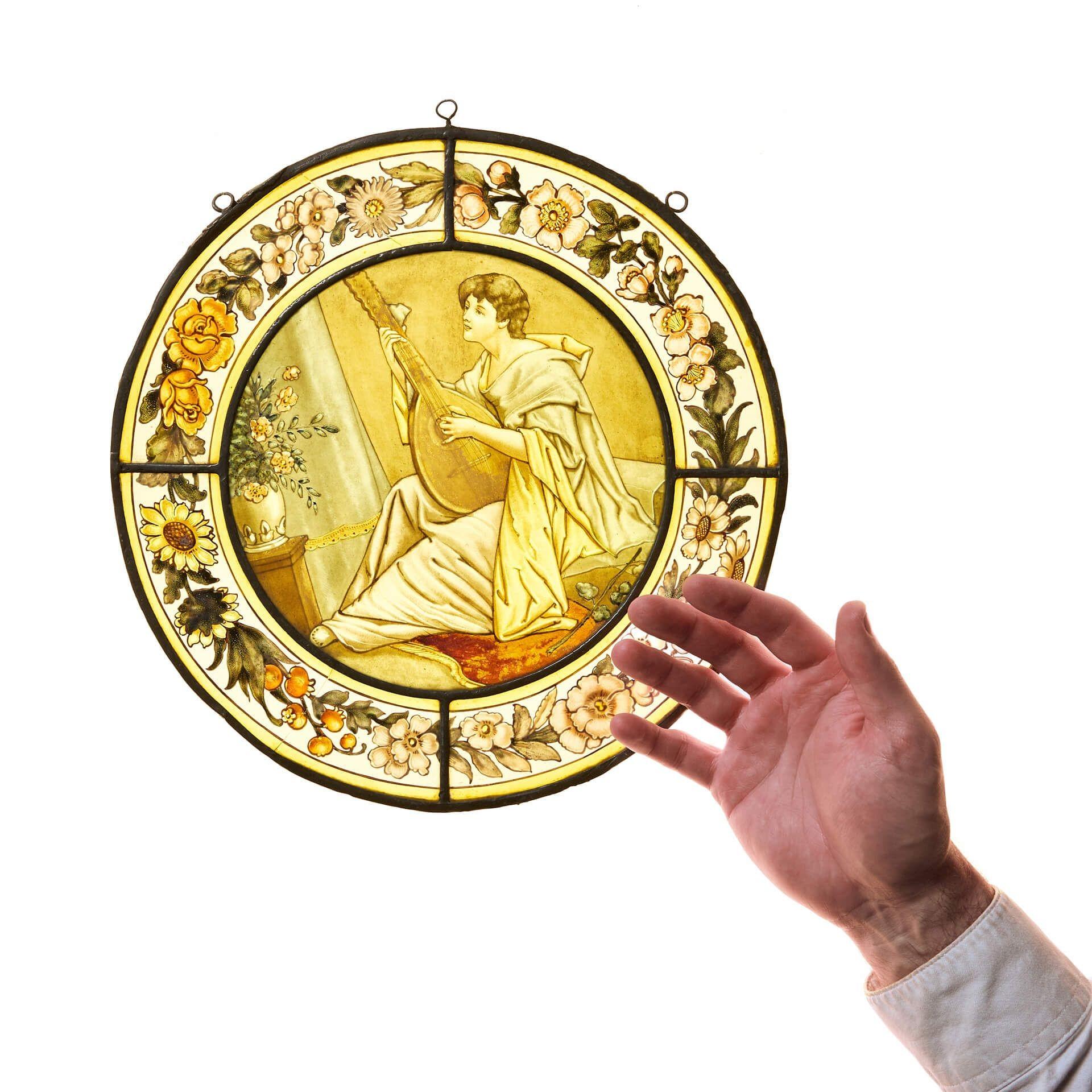 An antique Victorian stained glass window depicting a woman playing a lyre. Hand-painted stylised flowers with a vibrant yellow border surround the woman while she effortlessly plays the instrument sat in her pale blue and gold robes. A detailed