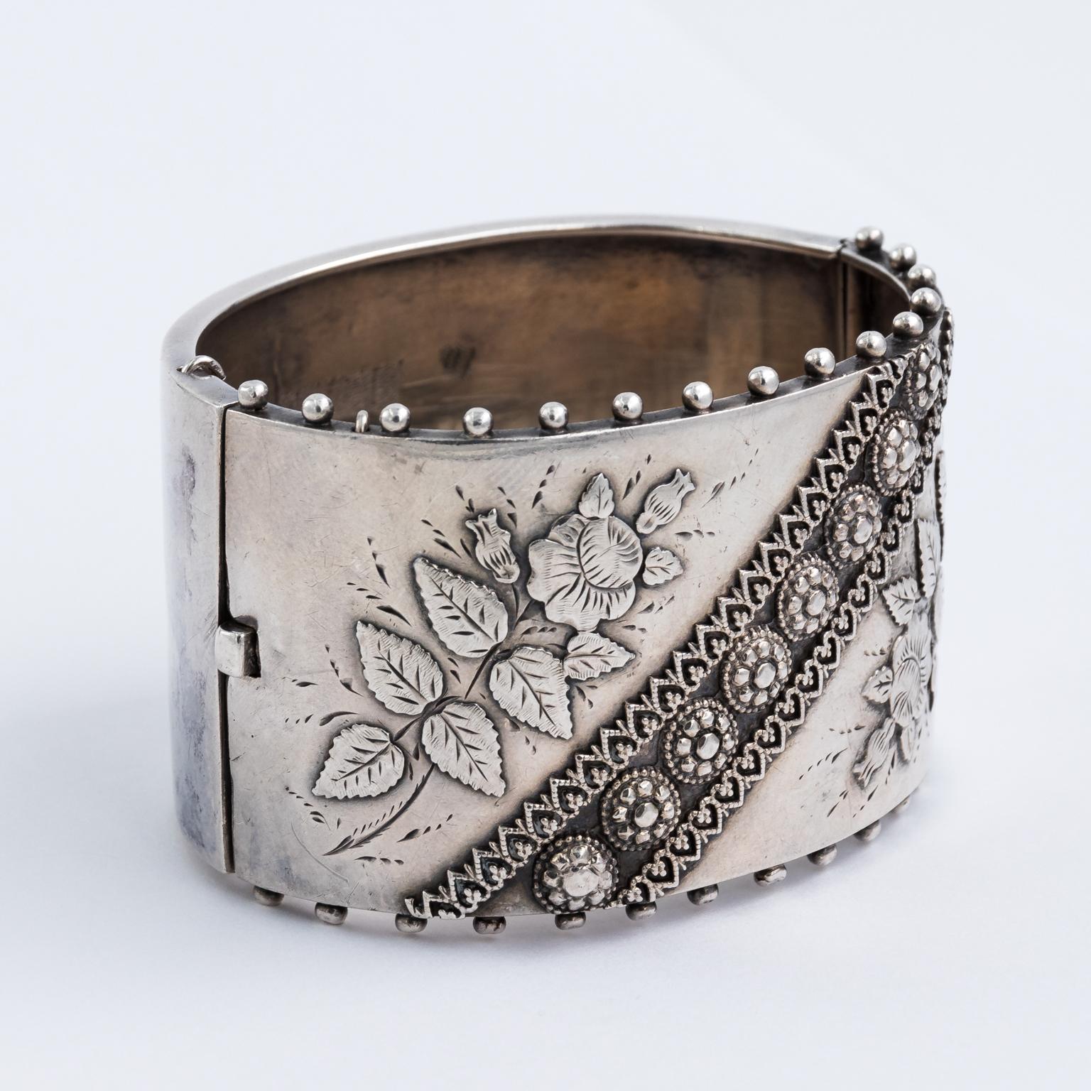 Circa 1880s English Victorian style silver bangle cuff bracelet with repousse Aesthetic Movement style floral motifs and linear trim. Hand marked in Sterling Silver with hinged clasp and safety chain.
