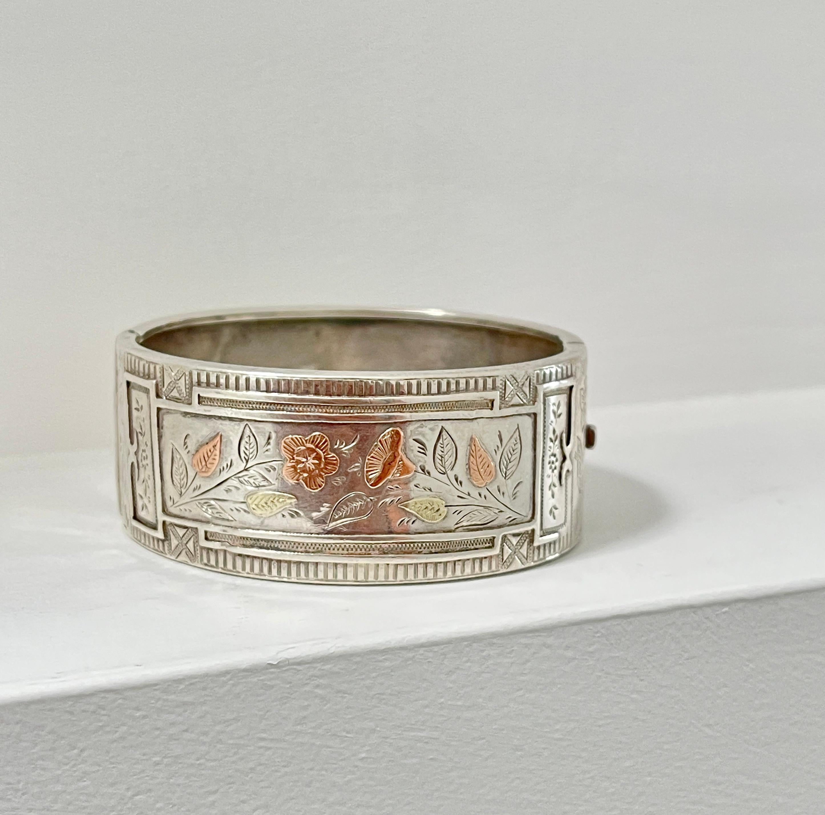 This beautiful Victorian bangle is 140 years old!

It is solid sterling silver and features a hand engraved flower and leaf design inlayed in yellow and rose gold.  The bangle was made in Birmingham in 1884 and bears these hallmarks plus the makers