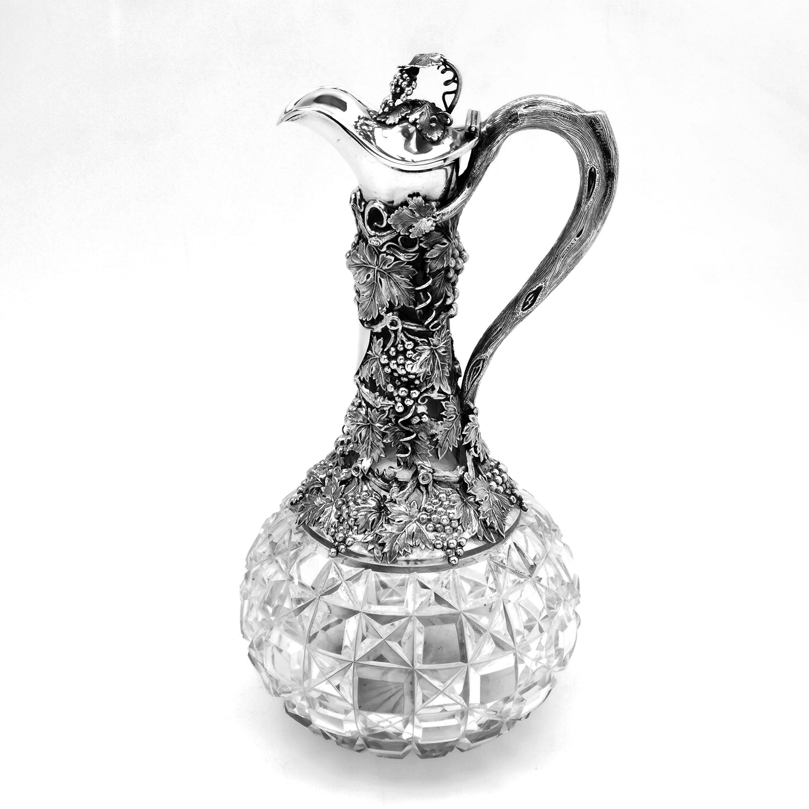 An antique Victorian sterling silver mounted claret jug with a cut glass body. The silver neck of the claret jug has an intricate grape vine design showing bunches of grapes and detailed leaves culminating in the handle of the jug designed to look