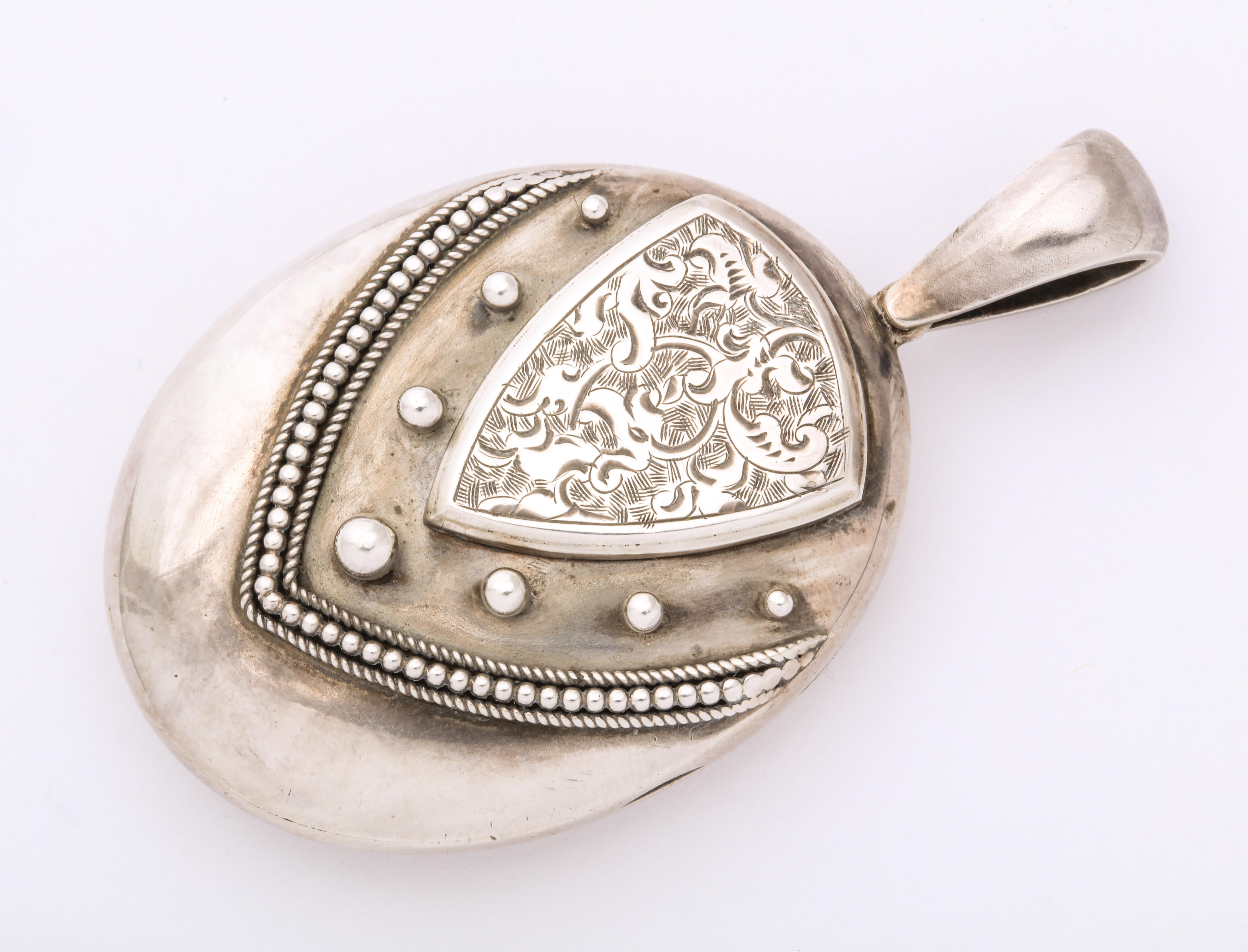 Contrasts of smooth and frilly engraving make an unusual shield design on this Victorian Silver Locket c. 1860-1880. The shield shape at top is covered with abstract vines atop a larger shield of raised silver graduated orbs, bordered by silver