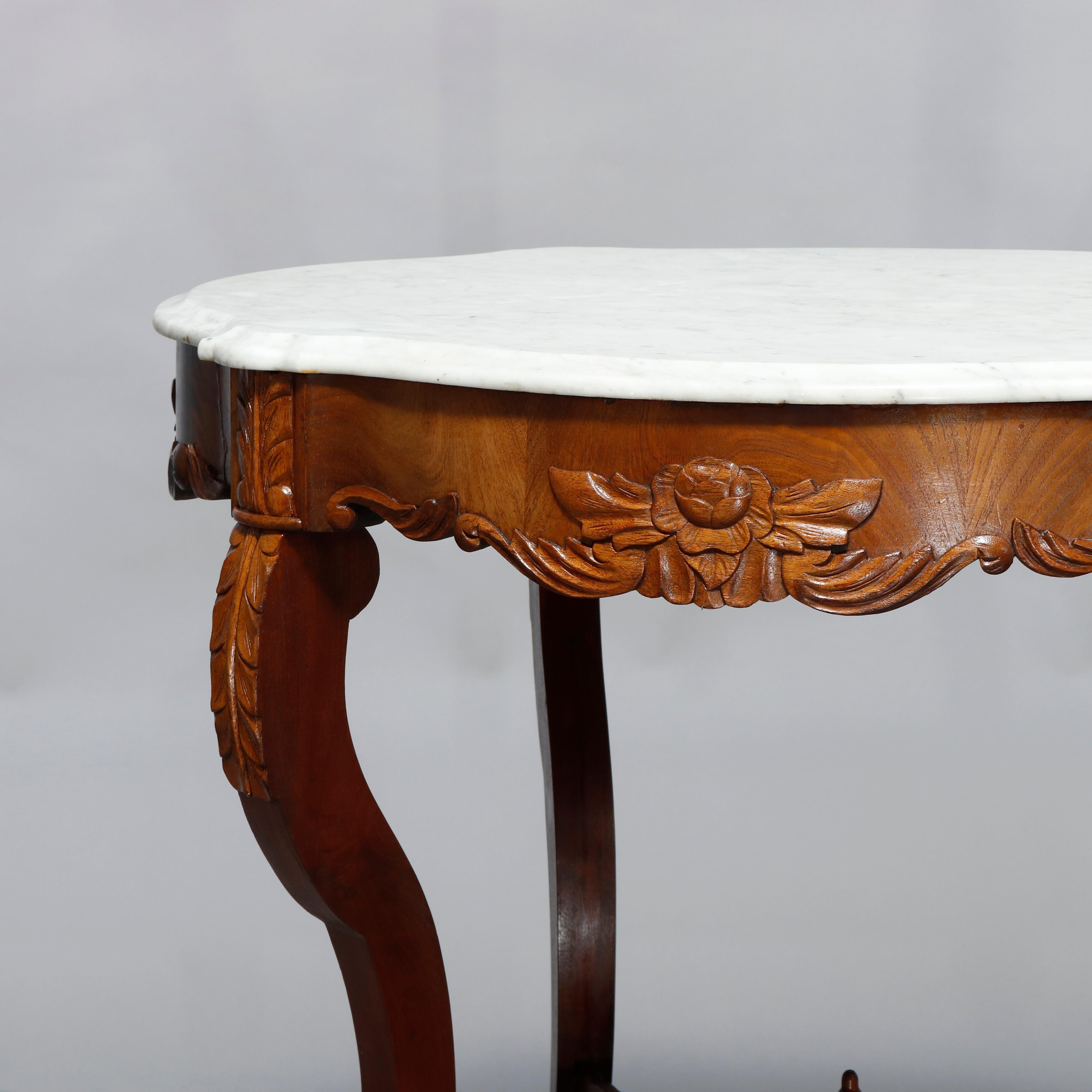 An antique Victorian turtle top lamp table offers shaped and beveled marble top over carved mahogany and walnut base having apron with foliate elements, raised on cabriole legs with scroll form stretcher having central urn form finial, circa