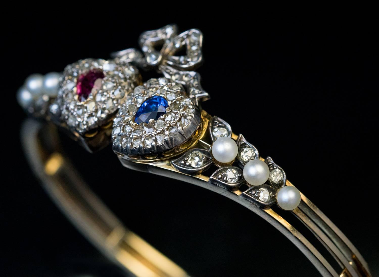 Circa 1880s

Un bracelet bangle ancien en or 14K est centré de deux cœurs en argent sertis d'un rubis en forme de poire et d'un saphir bleu (environ 0,30 ct et 0,35 ct) entourés de diamants taille rose. Les cœurs sont surmontés d'un nœud en diamants