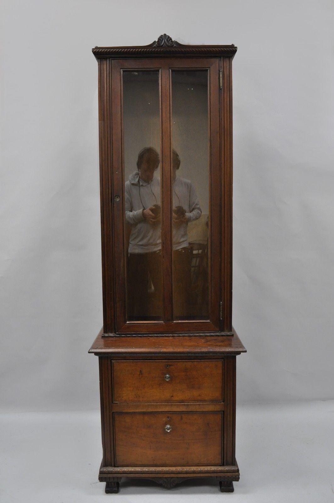Antique narrow two-piece walnut gun cabinet. Item features solid two-piece construction, nicely carved details, one swing door with glass, two drawers, attractive aged patina to wood, glass pulls, unlocked and locks not in working order. Upper