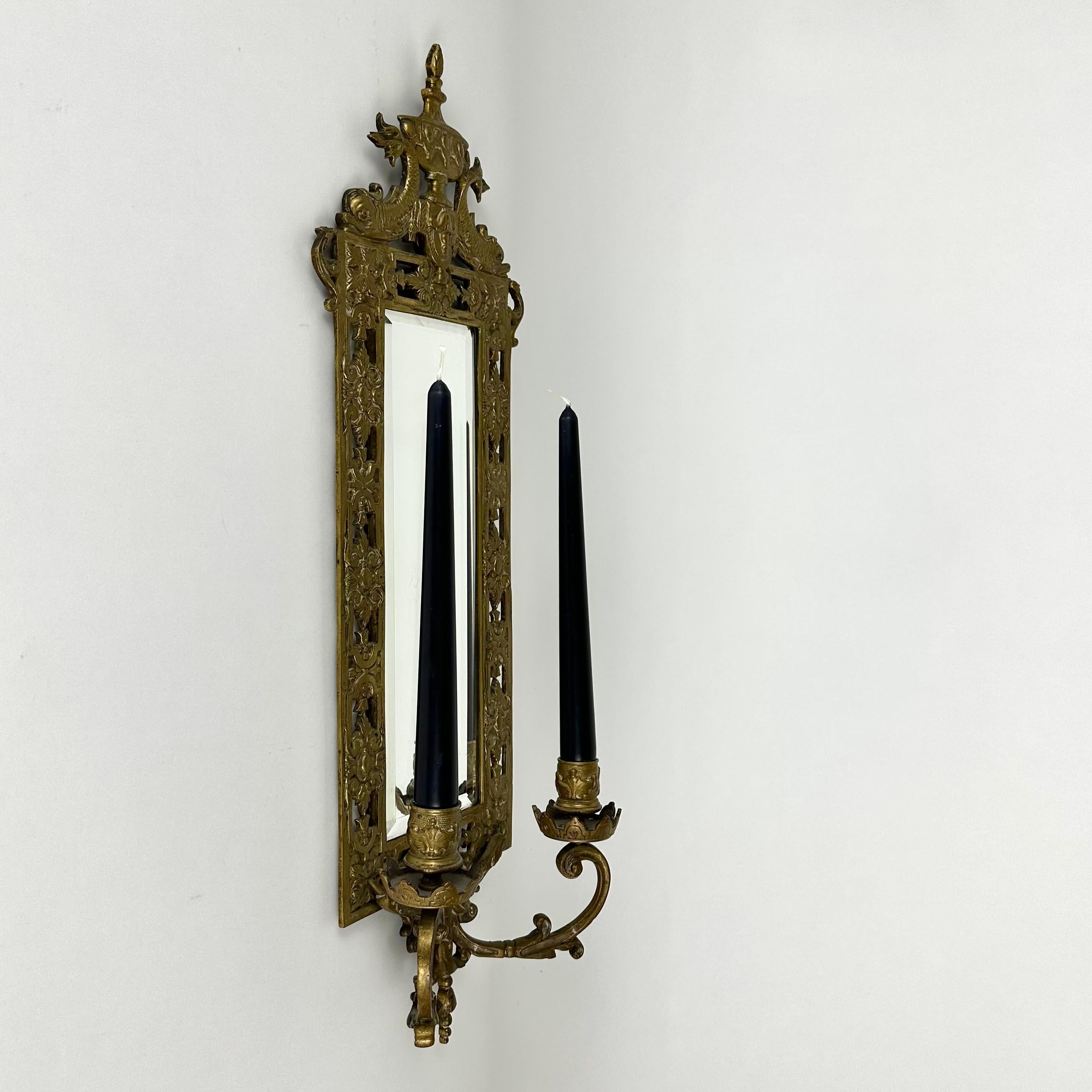 A classic mirrored sconce, rectangular in form. France, 1910s.

The top ornament features two classical dolphins.

Two arms at the bottom to hold taper candles.

The mirror is beveled, and has age spots in the original backing. The bronze has an