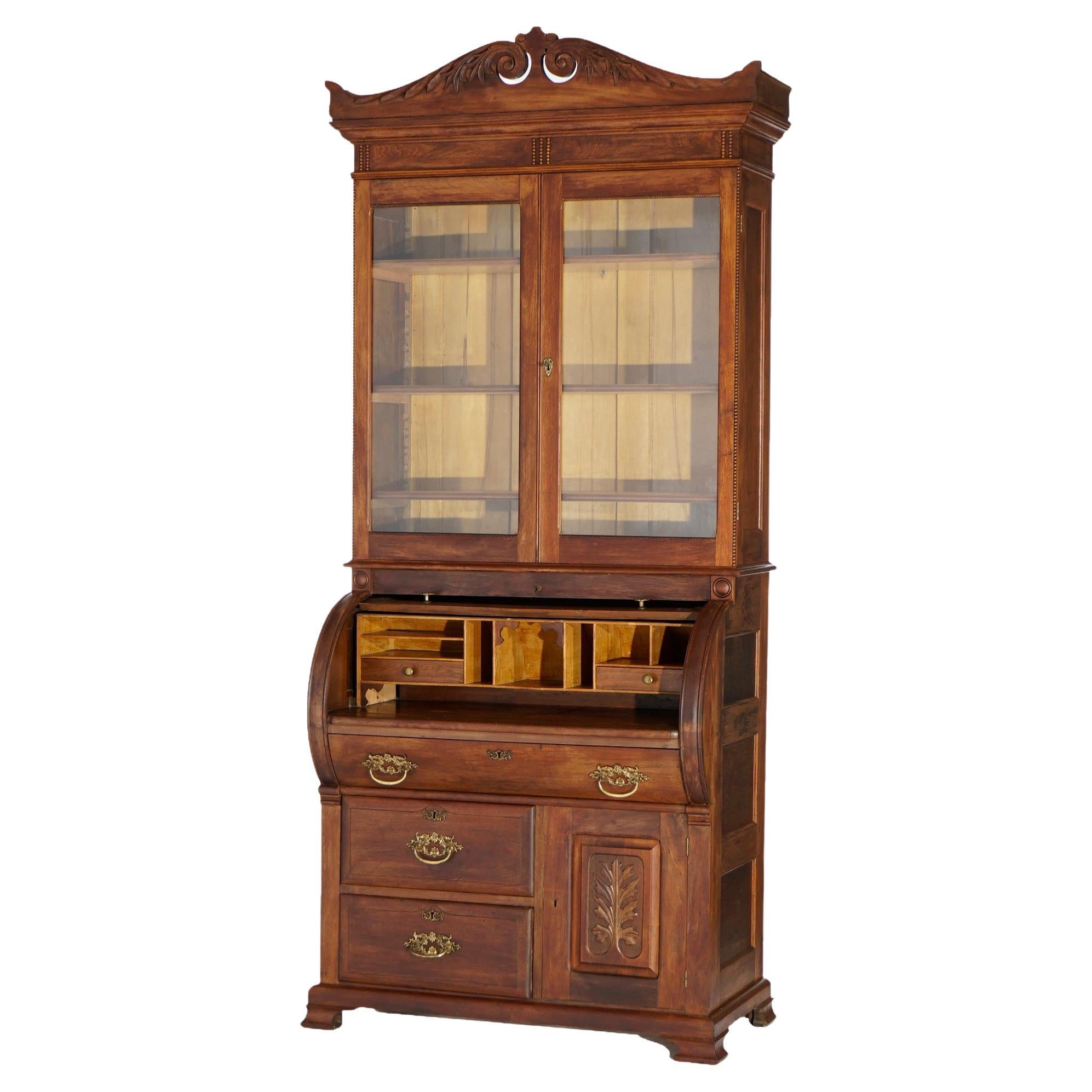 An antique secretary offers walnut and burl paneled construction with upper bookcase having broken arch crest over double glass door case seated on desk with cylinder opening to writing surface and storage compartments and lower drawers and cabinet