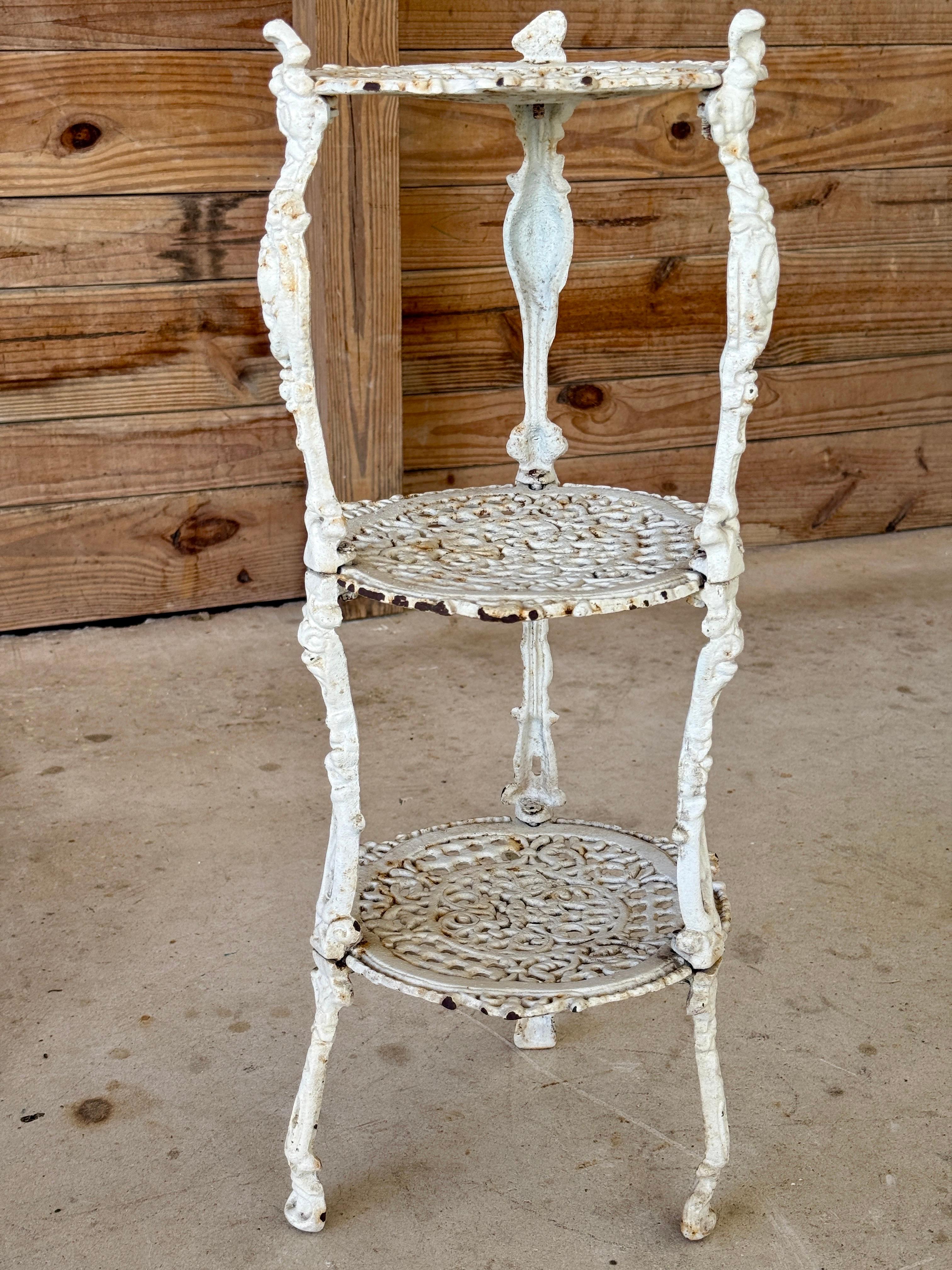 This charming antique three-tier cast iron plant stand features ornate scrollwork and classical detailing throughout, all finished in a beautifully aged white paint with natural chippy patina. Perfectly sized for a fern, bust, or accent lamp, it’s