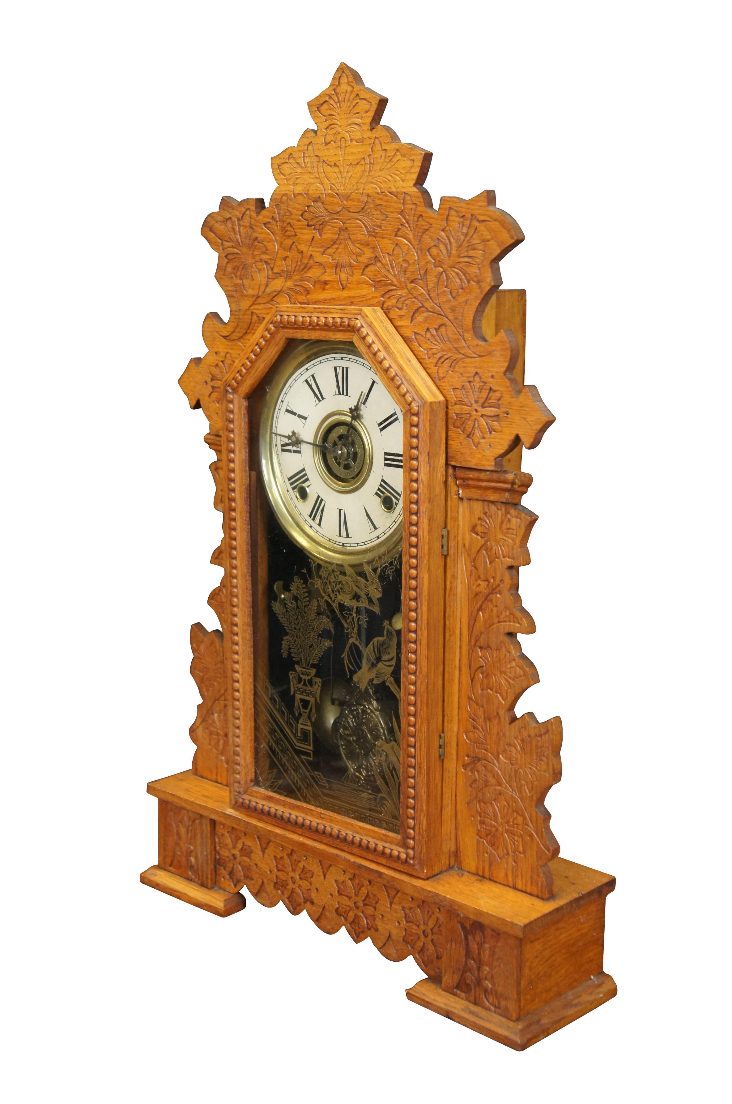 Late 19th - early 20th century William L. Gilbert key wound, 8 day, chiming gingerbread style mantel clock. Ornate oak case with a press carved ivy design and beaded trim; gold reverse painted glass front featuring a neoclassical vase and birds on a
