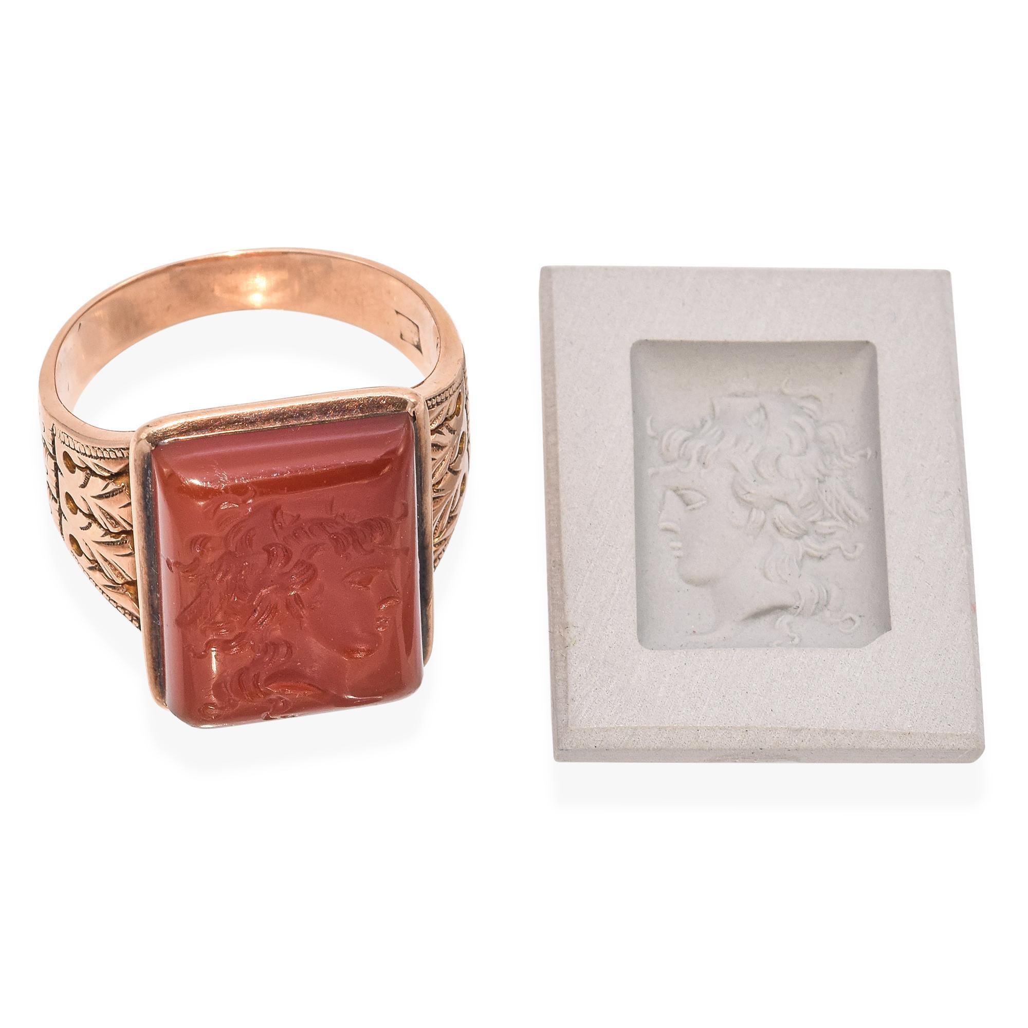Exuding timeless elegance, this striking ring features a rectangular Carnelian intricately carved with a rare double head. The stone is set in a warm, gold-toned band adorned with detailed leaf motifs on the sides. The ring will come with a clay