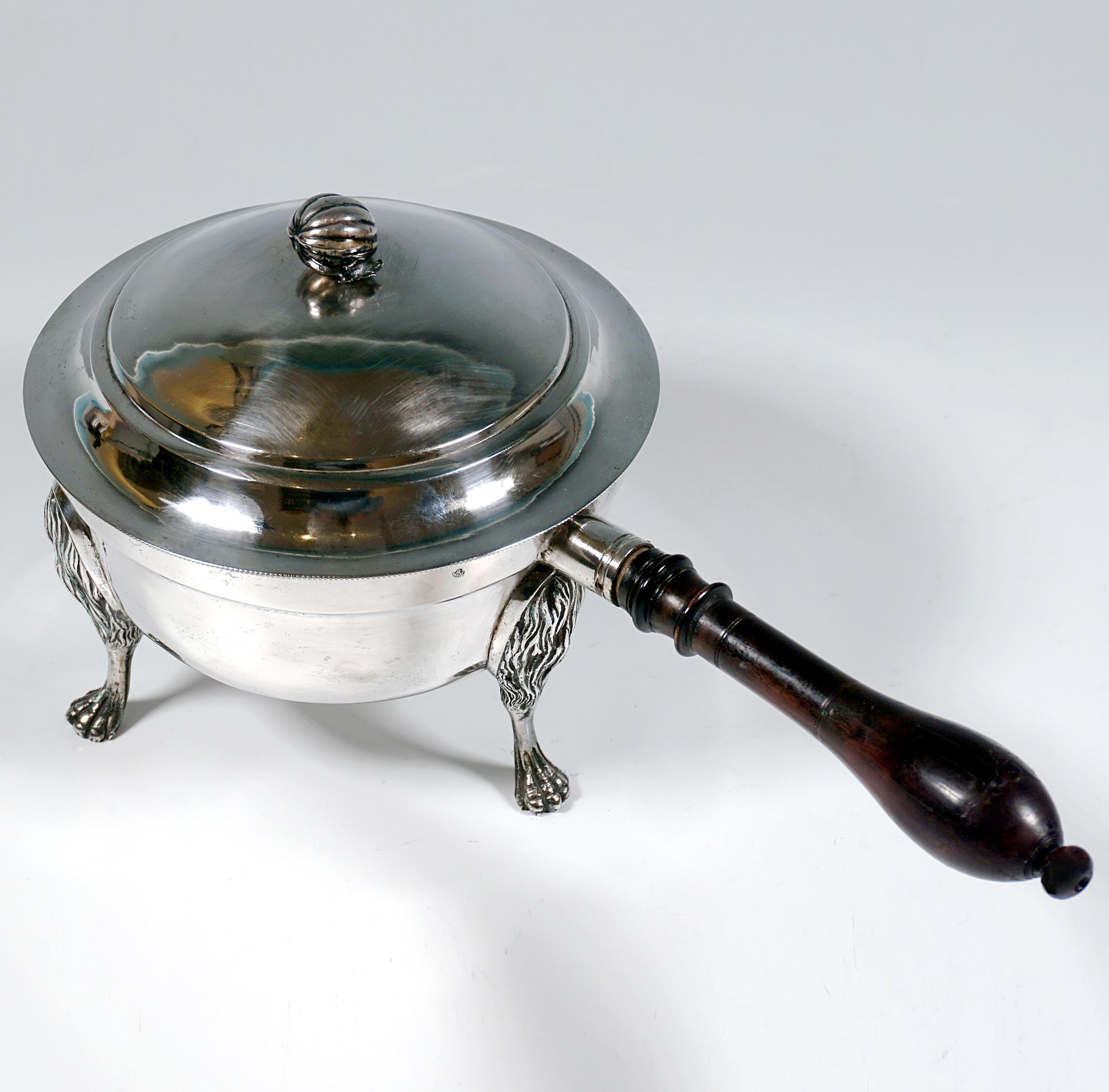 Cacerola de plata antigua, recipiente redondo y acampanado con paredes lisas y borde escalonado hacia fuera, de pie sobre tres patas de león con relieve de piel y patas esculpidas, montadas en el cuerpo del recipiente, con un asa de madera torneada