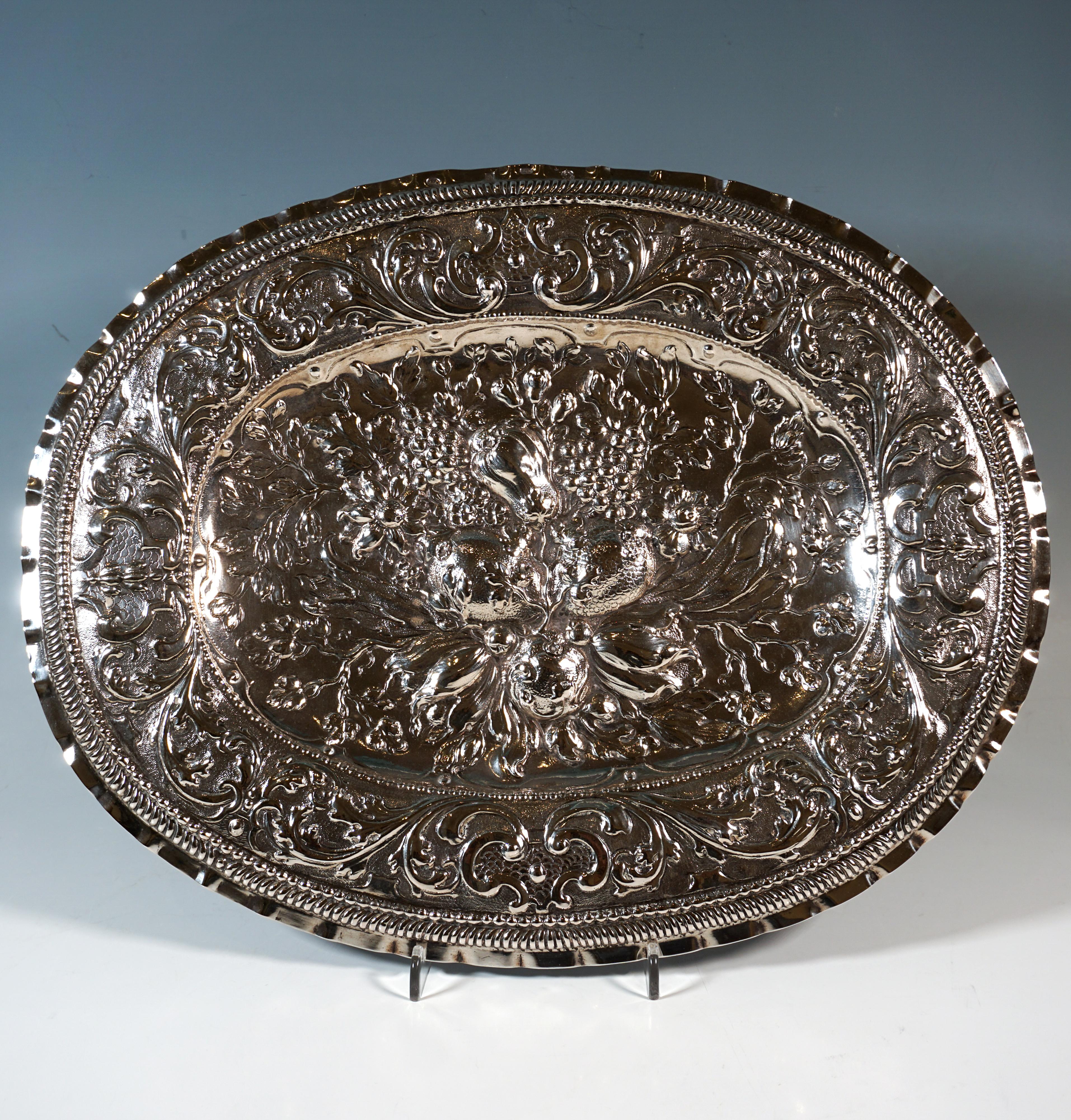 Piatto ovale in argento con specchio leggermente incassato e bordo a cupola, compensato da una delicata fascia di perle e da un arco in rilievo verso l'interno, nonché finemente sbalzato e inciso su tutta la superficie, centrato dalla forma di un