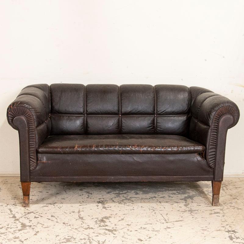 The rolled arms and back of this vintage brown leather sofa are accented by leather strips which adds to their visual appeal. There is age related wear to the dark brown leather which is more extensive to the seat cushion and arms, also separation
