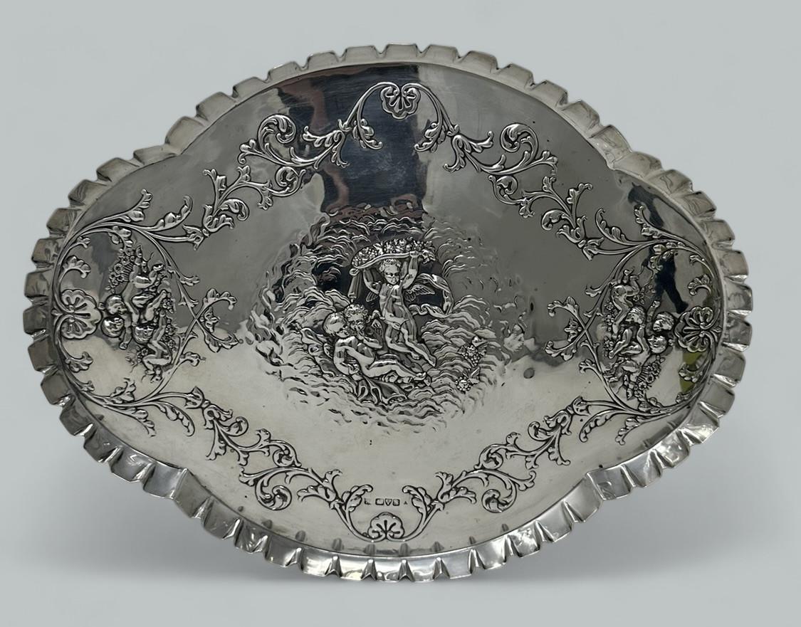 Elegante bandeja o tarjetero inglés de plata de ley, de contorno ovalado y buen tamaño y peso, con un detalle muy decorativo en el borde de la galería plisada. 

Todo ello repujado con flores, hojas y volutas, con elementos de decoración Art