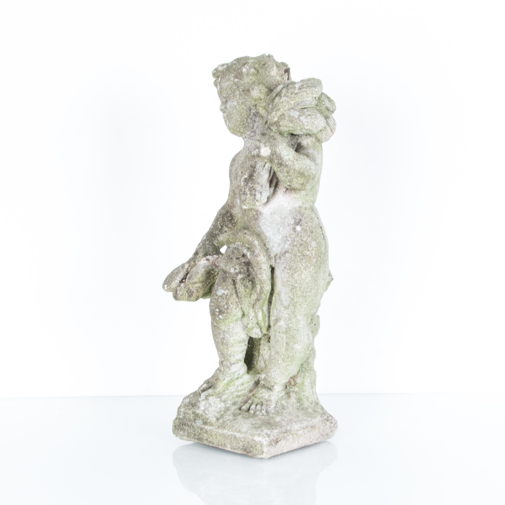 This concrete cherub statue was originally made in France, circa 1950. It holds a bushel of wheat over its shoulder, a symbol of natural abundance. The weathered grain of the concrete invests the piece with a Rococo softness, making this antique